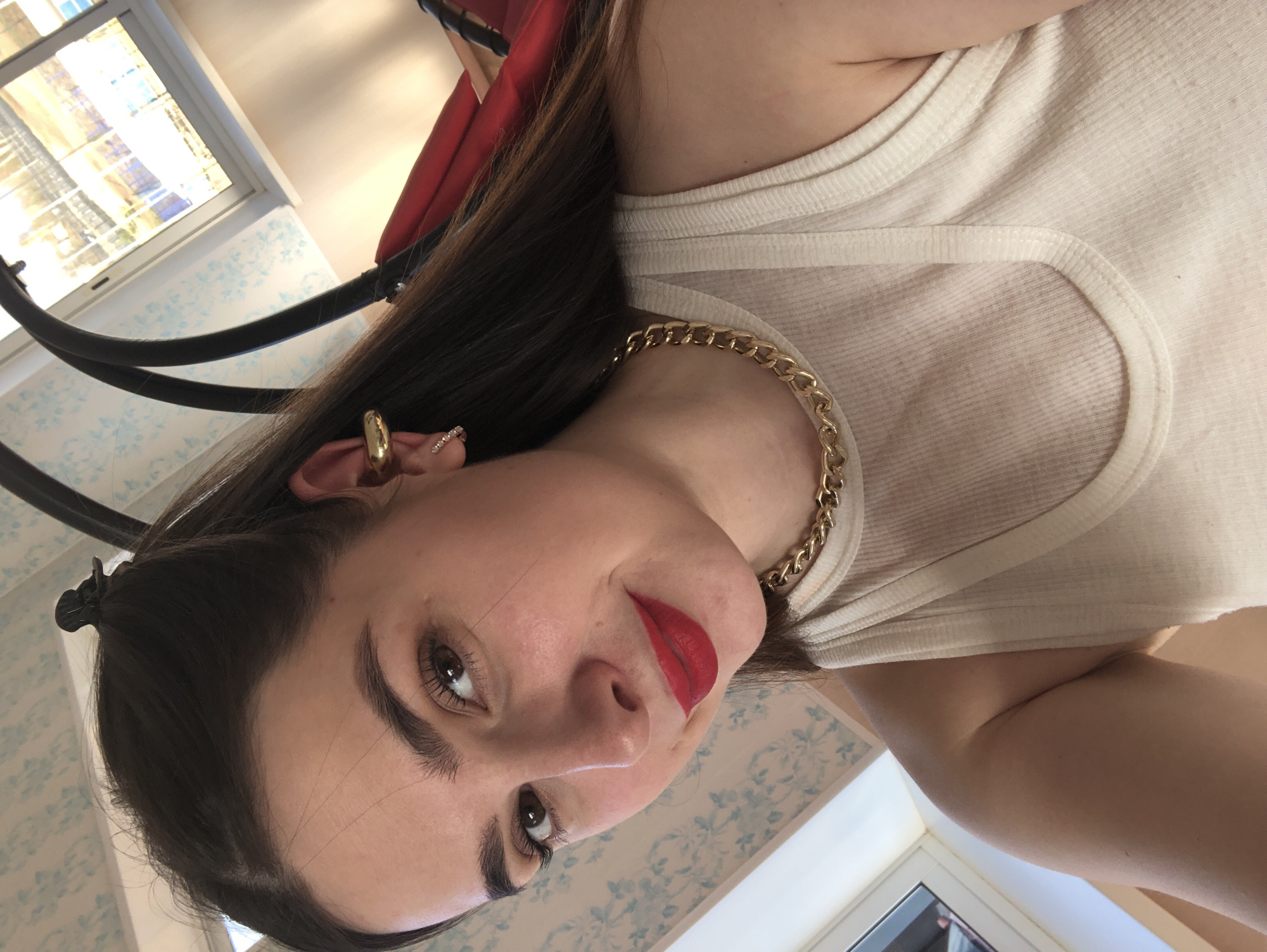 A medium close-up image, taken from a low and significantly tilted angle, features a young woman posing for a selfie. She has dark brown hair, which is partially pulled back with a small black clip visible at the crown of her head. Her dark eyebrows are well-defined, complementing her dark brown eyes, which show visible mascara on the lashes. She wears bright red lipstick, contrasting with her fair complexion.
She is dressed in a light-colored, likely cream or white, ribbed tank top. Around her neck, she wears a prominent chunky gold chain necklace. Her left ear is adorned with at least two pieces of jewelry: a large gold ear cuff or hoop on the upper cartilage and a smaller, sparkling stud or cuff below it. A small, sparkly ring is also visible on her left pinky finger. Her expression is calm with a hint of a subtle smile.
The background indicates an indoor setting during the daytime, with bright natural light entering through a window located in the upper left of the frame. The window features a grid pattern. Below it, a wall covered in light blue wallpaper with a subtle, intricate pattern is visible. Behind her, a dark, curved object, possibly part of a chair, a swing, or some other furniture, and a blurred red surface are also partially seen. No visible text is present in the image. The provided location context for the image is Noviy, Russia.