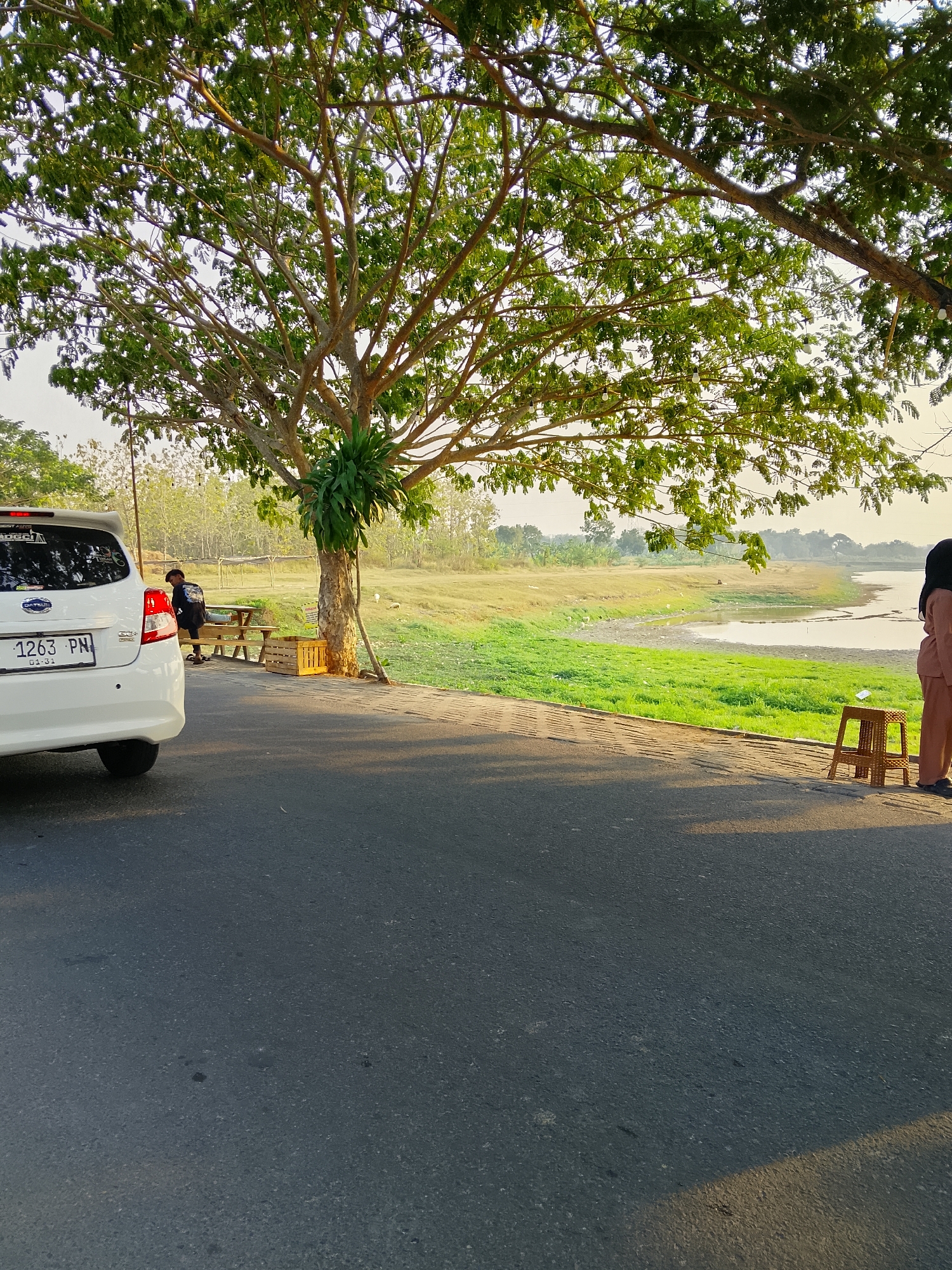In Ngadrin, Indonesia, this image captures a scene by a body of water. A white car is parked on the left side of the frame. To the right of the car, a young person sits on a wooden picnic bench under a large tree. A small wooden crate is placed beside the tree. Further to the right, a person wearing a headscarf and a brown outfit stands near a small, woven stool. The environment features a road in the foreground, with a grassy area and the water body extending into the background. Trees and vegetation surround the water, with a hazy sky above. The lighting suggests it is either early morning or late afternoon, with the sun casting shadows.