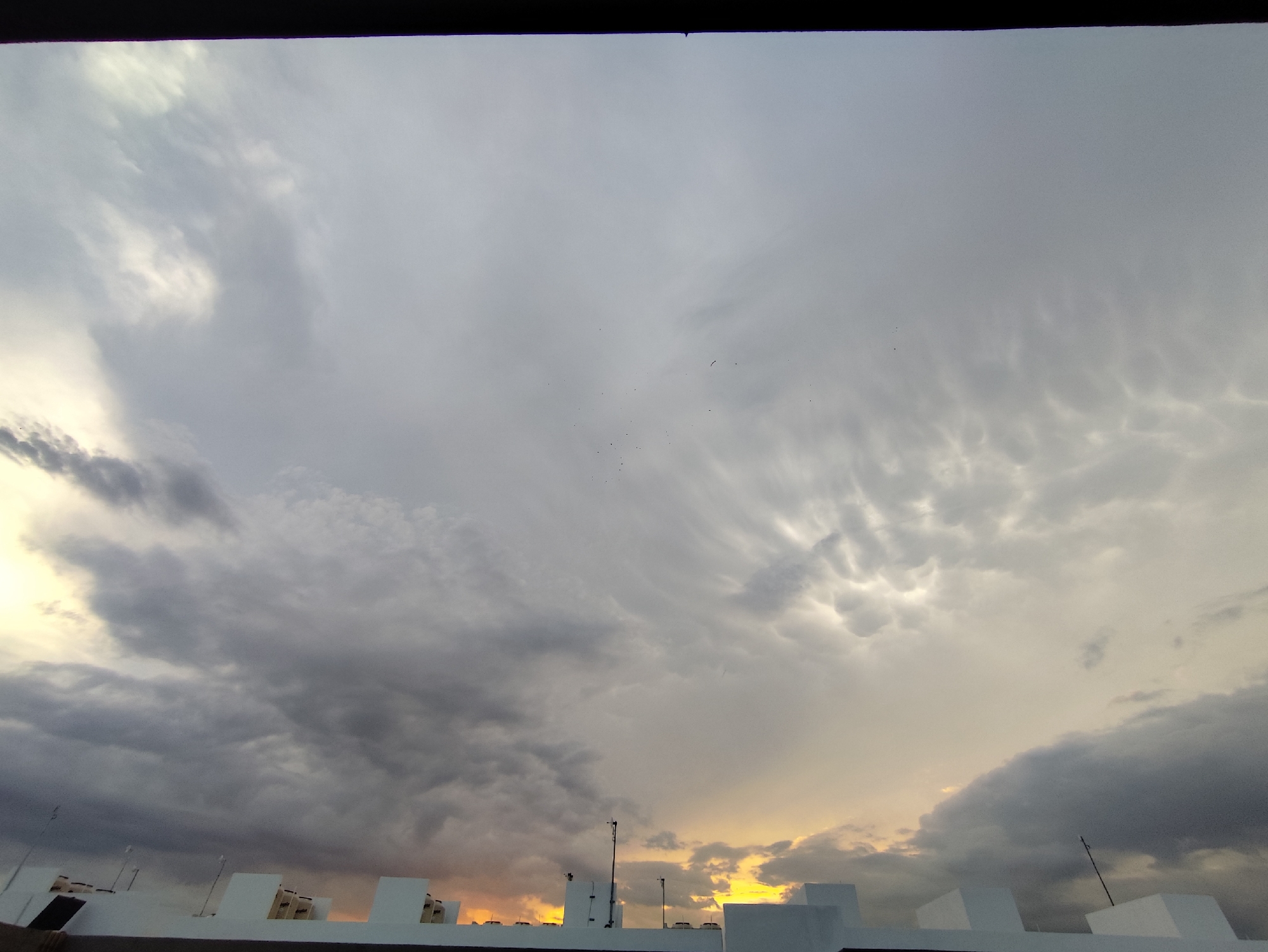 A wide shot captures a cloudy sky over Playa del Carmen, Mexico. In the foreground, a white rooftop with multiple square and rectangular protuberances is visible. Several small antenna-like structures are scattered across the roof. Behind the rooftop, a yellow-orange glow suggests either sunrise or sunset. The sky is predominantly covered with grey and white clouds, displaying varying textures and densities. A few tiny, indistinct objects that may be birds are seen higher in the sky. The overall impression is of an overcast day with some potential for dramatic lighting effects.