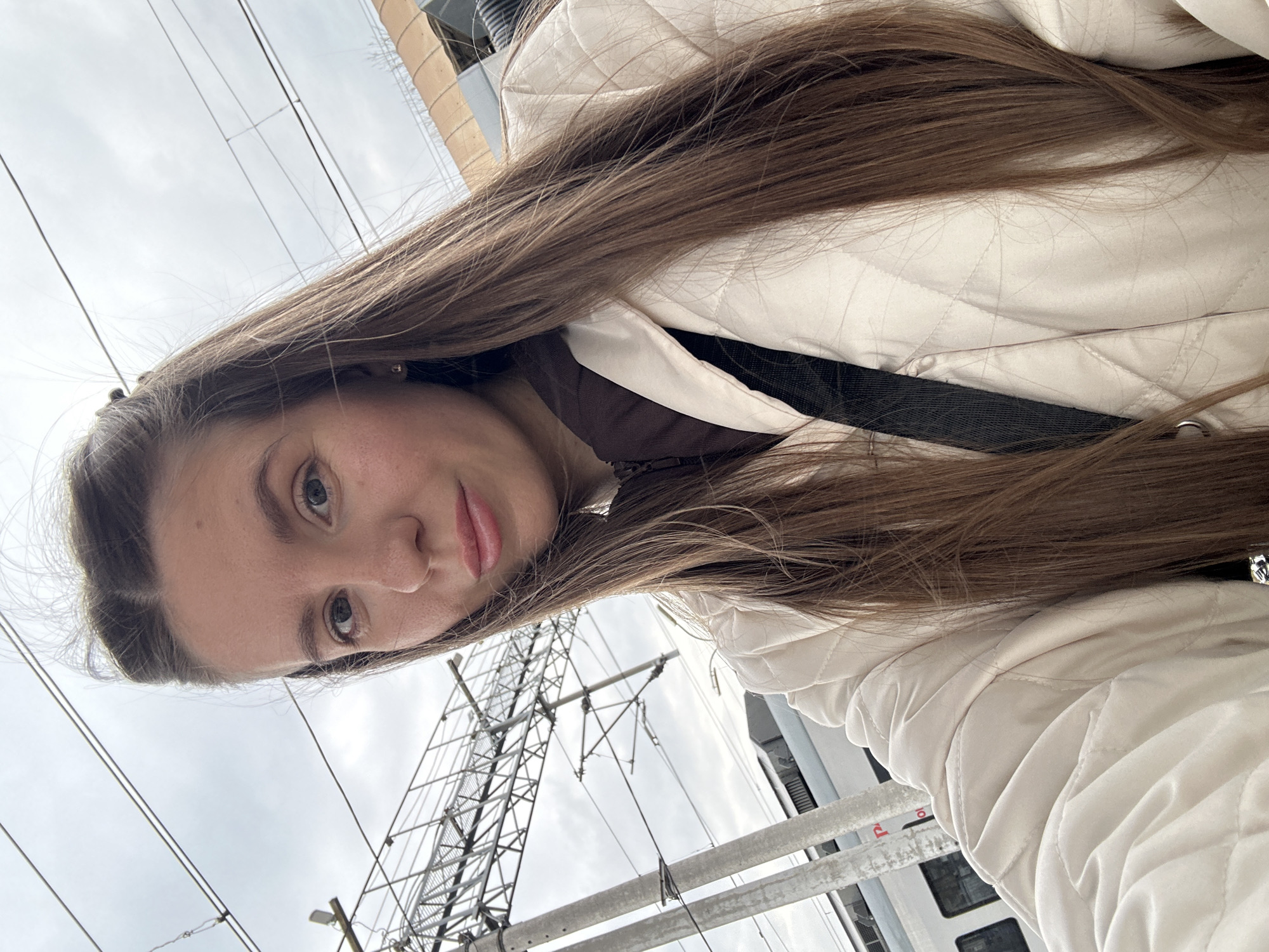 A young woman is captured in a close-up selfie, looking directly at the camera with a neutral expression. Her long, straight brown hair cascades around her face and over her shoulders. She is wearing a cream-colored, diamond-quilted jacket over a darker top. The image is taken from a slightly upward angle.
The background shows an overcast sky, indicating cloudy daytime conditions. Numerous overhead power lines and cables crisscross the frame, supported by utility poles and substantial metal lattice structures, strongly suggesting an industrial or railway environment. The side of a train car is partially visible in the lower right background. Faint, indistinct red text on a white surface can be seen, likely on the train or a nearby sign. The scene is set outdoors, consistent with a location near train tracks in Klin, Russia.