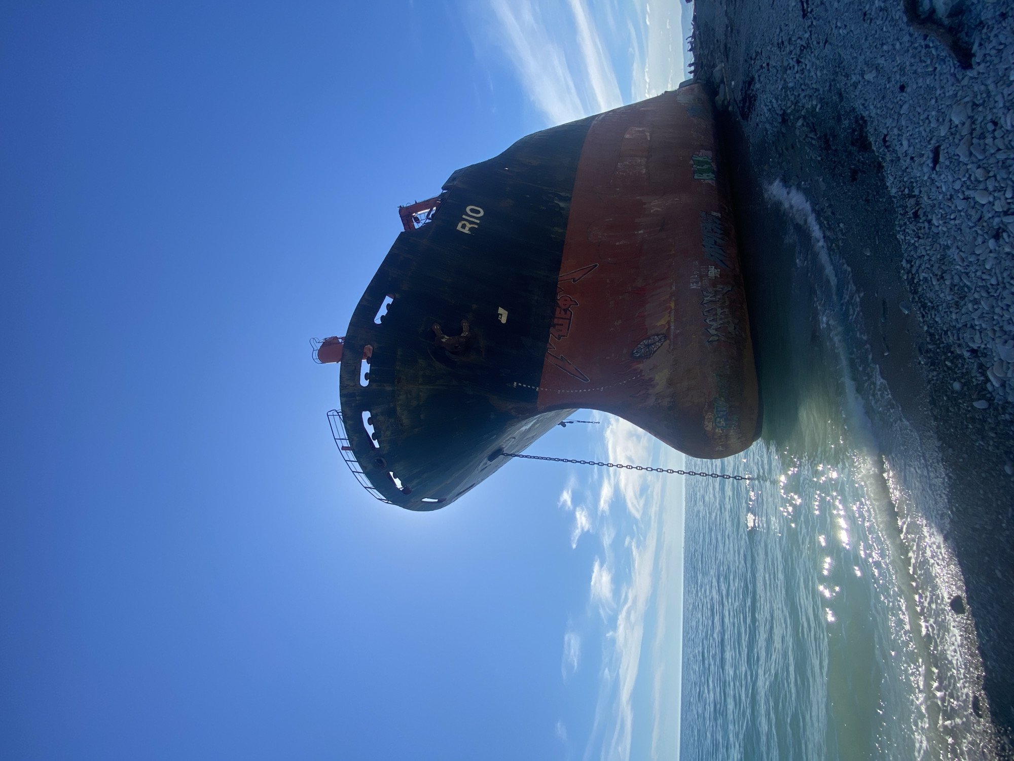 This media file captures a majestic yet derelict cargo ship, named "RIO," beached on a pebble shore under a bright, clear sky. The ship's bow is prominently displayed, facing towards the viewer and slightly to the left. The hull features a dark upper section and a rusty red lower part, with noticeable wear, patches of rust, and various instances of graffiti, particularly on the red portion. The name "RIO" is clearly legible in white capital letters on the ship's upper bow. An anchor chain extends from the bow into the calm, sparkling blue-green water, which gently meets the dark, pebbled beach on the right side of the frame.
The sky is a vivid blue, adorned with scattered, wispy white clouds that suggest a serene, sunny day. Bright sunlight reflects intensely off the water's surface, indicating either mid-morning or early afternoon. The overall setting implies a sense of abandonment and the enduring presence of the massive vessel on the shoreline. No people are visible in the image. This scene is located at Kabardinka, Russia, where the ship has evidently been grounded for an extended period.