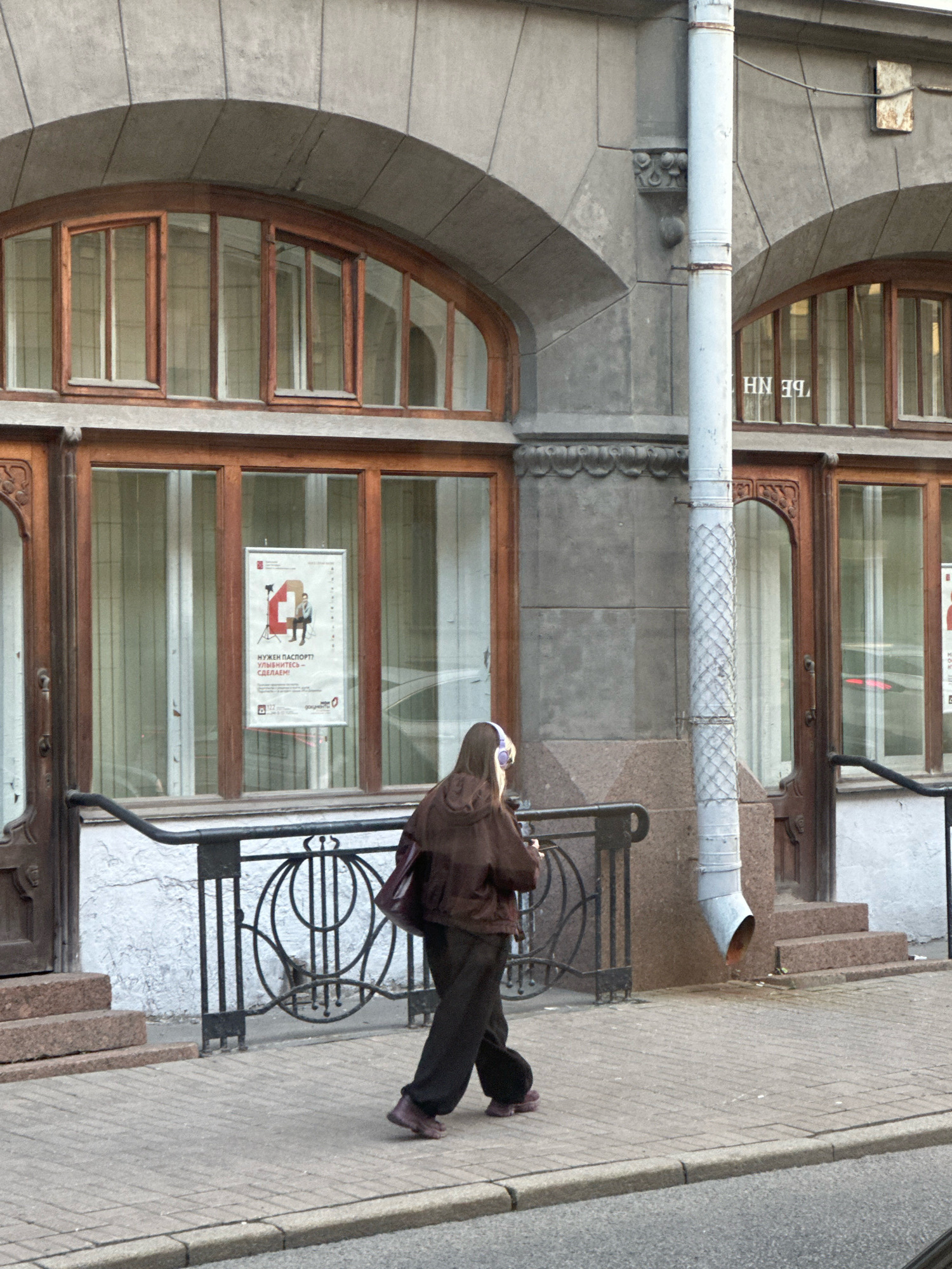 This image depicts a street scene in Saint Petersburg, Russia, focusing on the facade of a building and a pedestrian. The building features large arched windows with wooden frames, and ornate decorative elements above the windows. A light-colored downspout runs vertically down the building's exterior. A poster is displayed on one of the windows. A person wearing a brown hooded jacket and dark pants is walking on the sidewalk in front of the building. The person has headphones on. The sidewalk is paved with bricks, and a metal railing with decorative elements runs along the front of the building. Steps made of granite lead up to the building's entrance. The overall atmosphere is that of an urban environment in Saint Petersburg.