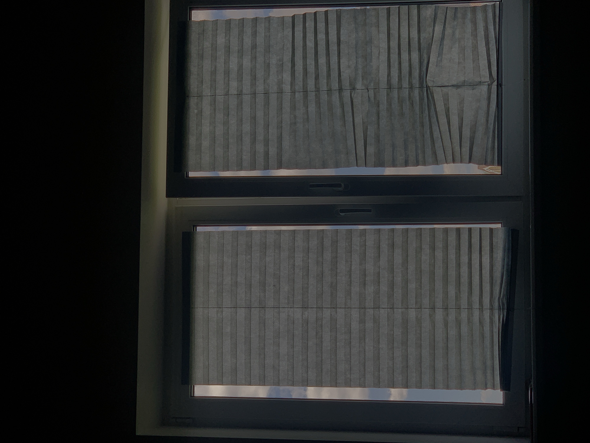 The image depicts two closed windows with pleated blinds. The window on the left is fully covered by the blinds, while the blinds on the right window are partially open at the bottom, revealing a sliver of the sky and clouds. The overall scene suggests an indoor perspective looking out. There is no information within the image to suggest a specific location in Minsk, Belarus, or to identify any people, actions, or interactions. The environment appears to be a room interior.