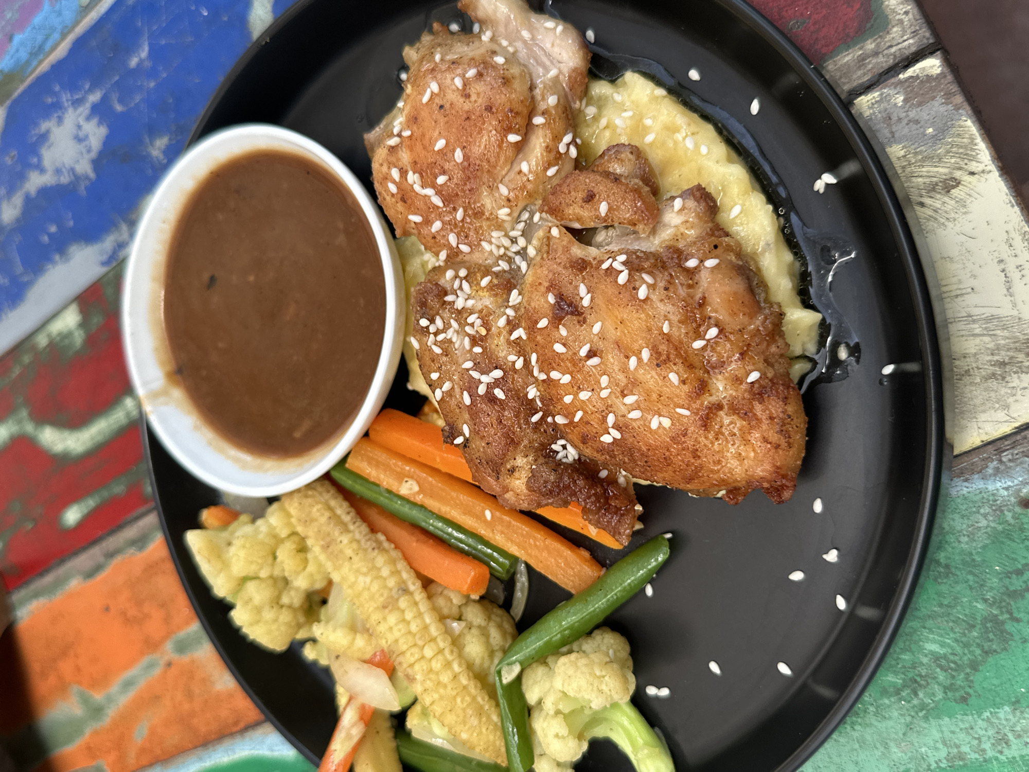 A black plate holds a meal of three pieces of fried chicken seasoned with sesame seeds, served over mashed potatoes and accompanied by a side of mixed vegetables and a small bowl of brown sauce. The vegetables include cauliflower florets, baby corn, carrots, and green beans. The plate is set on a colorful, rustic wooden table with a distressed finish. The lighting is natural and diffused.