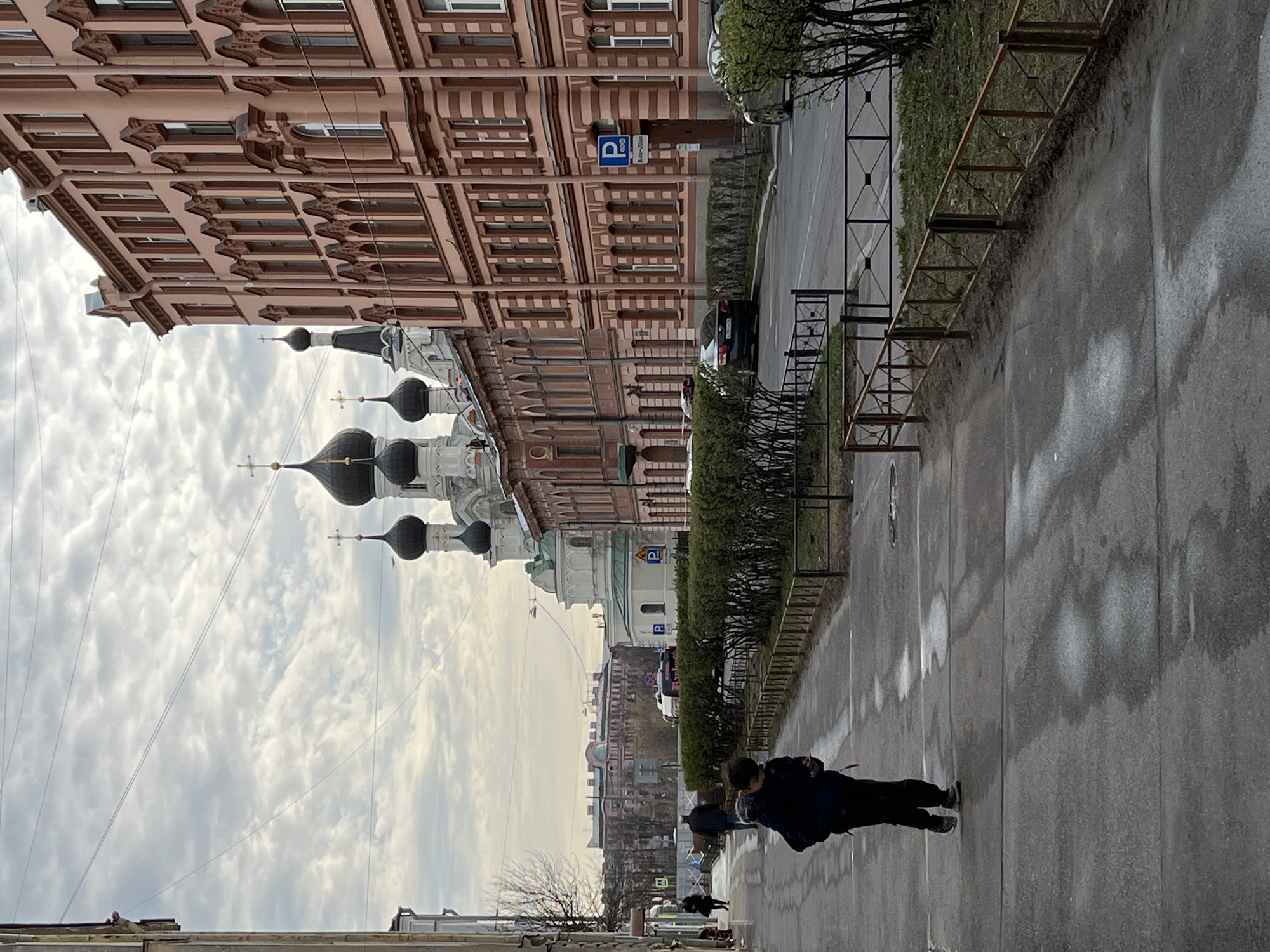 The image is a street scene in Saint Petersburg, Russia. The photo shows a pedestrian walkway along a street. The prominent features include the domes of a Russian Orthodox church. There are several people walking on the pedestrian walkway. Buildings line the street, with notable architectural details. A parked car is visible on the side of the street. The sky is overcast with clouds. A metal fence separates the walkway from a green area. The street appears to be wet, possibly from rain. There are trees with bare branches in the background. A parking sign is attached to one of the buildings.