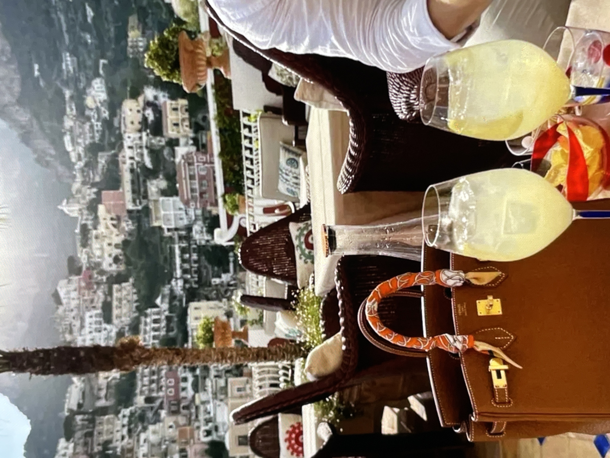 The image captures a luxurious outdoor seating area with a stunning panoramic view of a densely populated hillside town with white and pastel-colored buildings cascading down to the water. The foreground features a rich brown Hermes Birkin bag adorned with an orange and white silk scarf tied around its handles. Next to the bag are two glasses of a pale yellow beverage, likely a cocktail or spritzer, filled with ice and garnished with lemon slices. A small bowl filled with potato chips is also visible. In the background, wicker chairs and tables are set up for dining, suggesting a restaurant or private terrace. A tall palm tree stands to the left, and a glimpse of a person in a white shirt is visible on the right. The overall setting evokes a high-end vacation destination. There is no information about the city of Yalga, Russia, in this image.