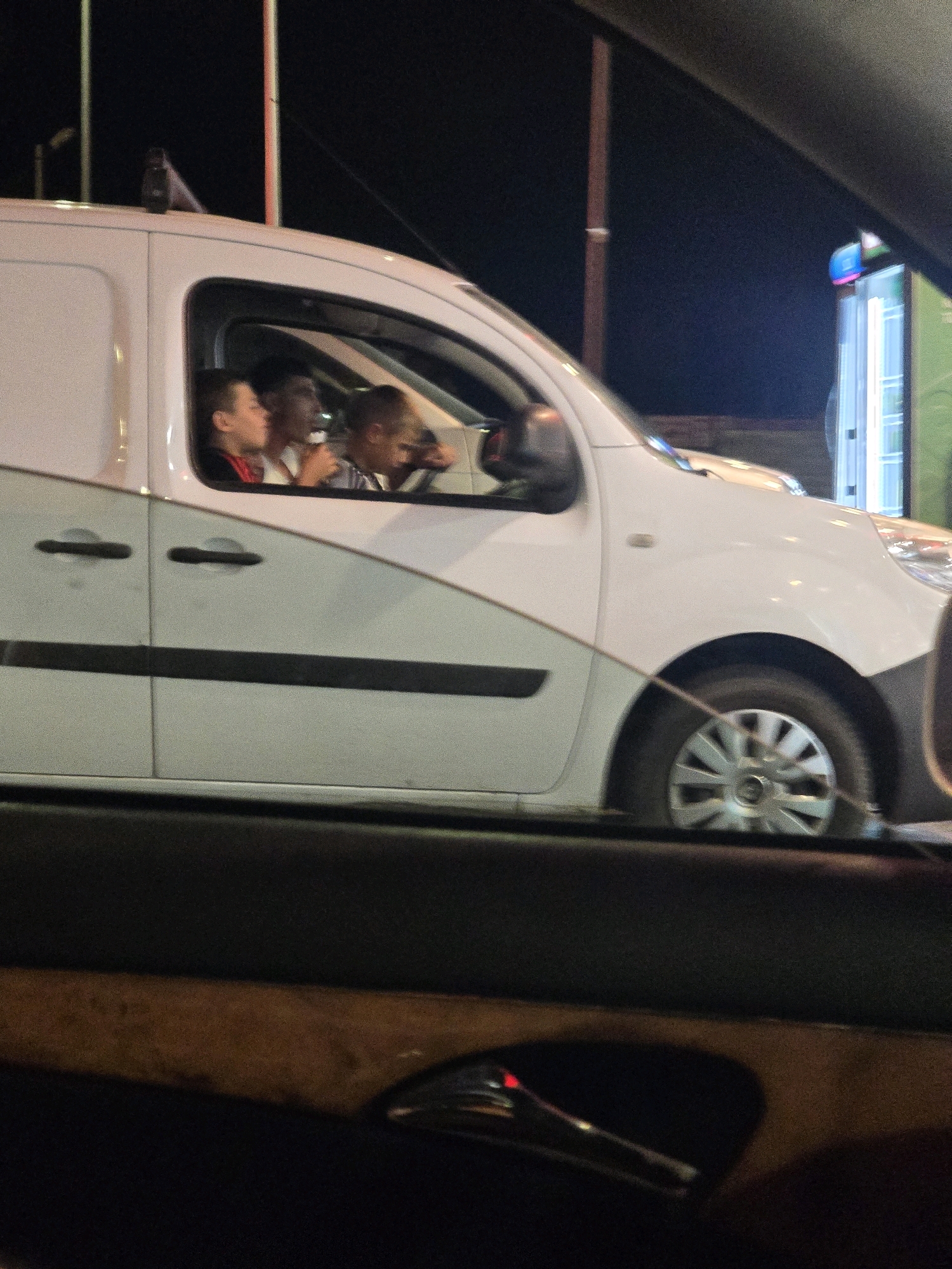 The image shows a white van at night. Three young individuals are visible inside the van through the passenger window. One individual appears to be driving. The van is parked near a brightly lit structure, possibly a storefront or a building with illuminated signage, which emits a blue and pink light. In the foreground, the dashboard and part of the interior of another vehicle are visible, suggesting the photo was taken from inside a car. The overall scene is dark, consistent with nighttime conditions.