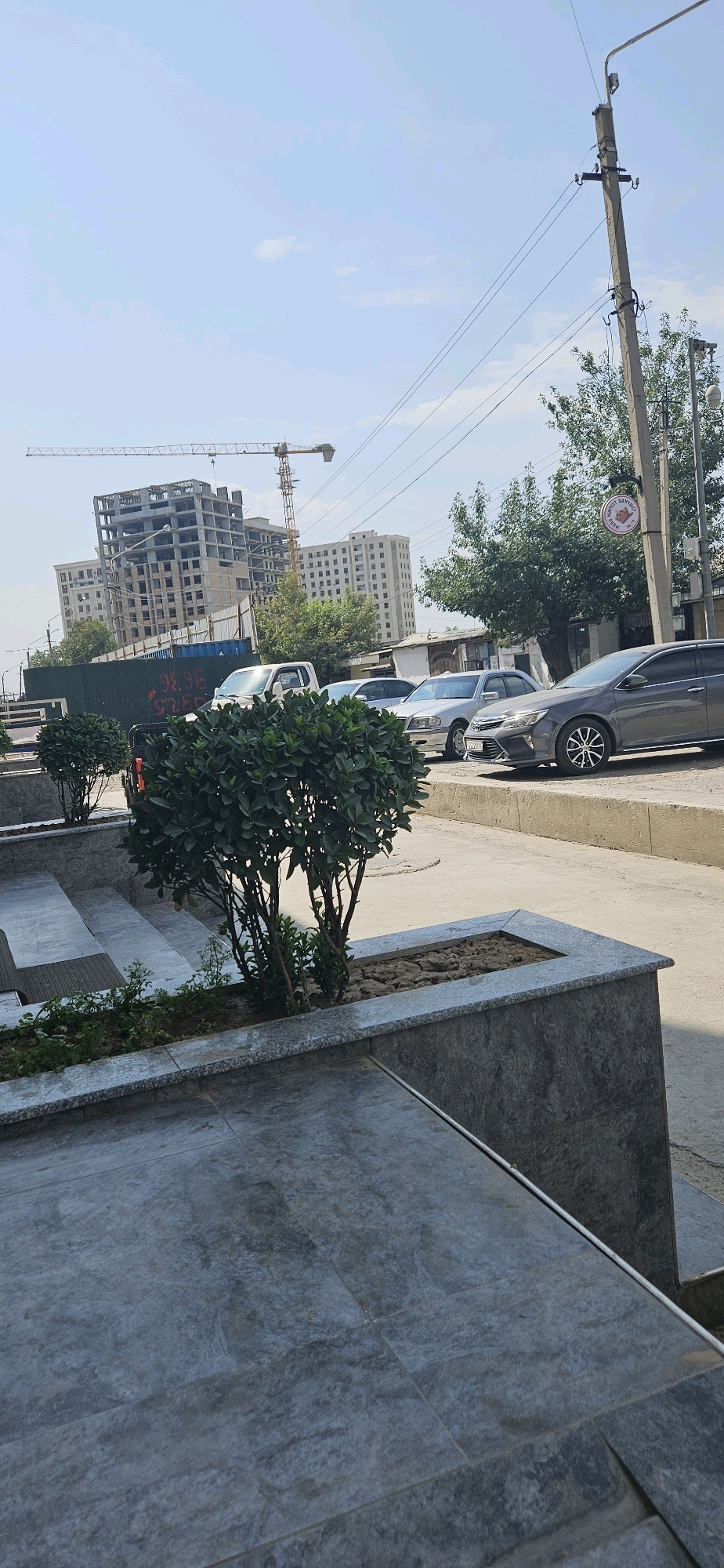 This image shows a street scene in Dushanbe, Tajikistan, with a focus on construction and urban development. In the background, a tall tower crane is visible, alongside unfinished multi-story buildings, indicating an active construction site. Several cars are parked along the side of the road, including a dark grey sedan and a silver sedan. A power pole with electrical wires runs along the right side of the image. In the foreground, there are tiered planters with greenery and stone steps leading up to a higher level. The sky is clear with a few scattered clouds. The overall impression is one of an urban environment undergoing modernization.