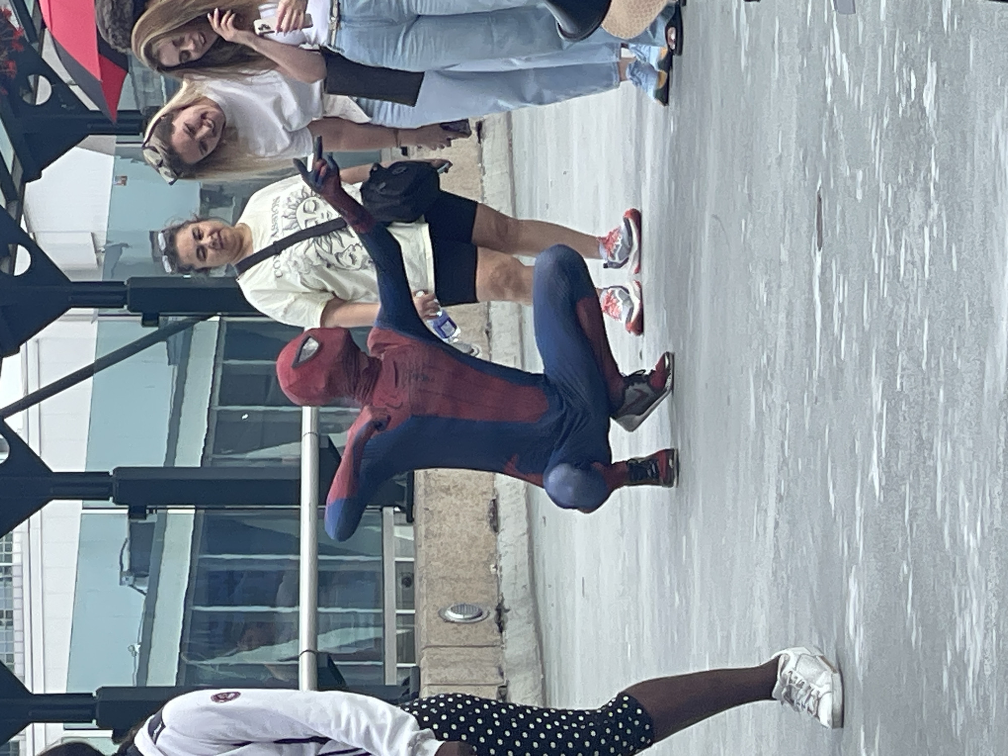 The image depicts a person in a Spider-Man costume crouching on a paved outdoor surface, likely a pedestrian walkway or plaza. The Spider-Man figure is in a dynamic pose, with one arm extended forward and the other bent. In the background, several people are visible. To the left, a person in a white hoodie and black and white polka dot pants is walking away. To the right, three women are standing and observing. One woman is wearing a cream-colored t-shirt, another a white t-shirt, and the third a white top with blue jeans. A red and black umbrella is partially visible in the upper right corner. The environment appears to be urban, with a building structure with glass windows in the background. The lighting suggests it is daytime. The scene is consistent with a public space in downtown Toronto, Canada, possibly near a transit hub or entertainment district where street performers might be present.