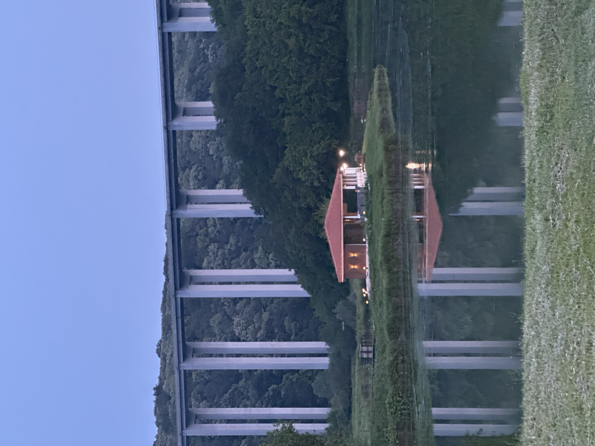 A tranquil twilight scene in Pisanets, Bulgaria, features a multi-pier concrete viaduct bridge spanning a densely forested valley under a clear, light blue sky. In the midground, a small, single-story building with a red-tiled roof and illuminated wall sconces rests on a grassy peninsula extending into a calm body of water. The water reflects the bridge's vertical piers and the building's warm lights. A small, dark platform structure is visible on the water's edge to the left of the building. The immediate foreground consists of a strip of mown green grass. No people or animals are visible.