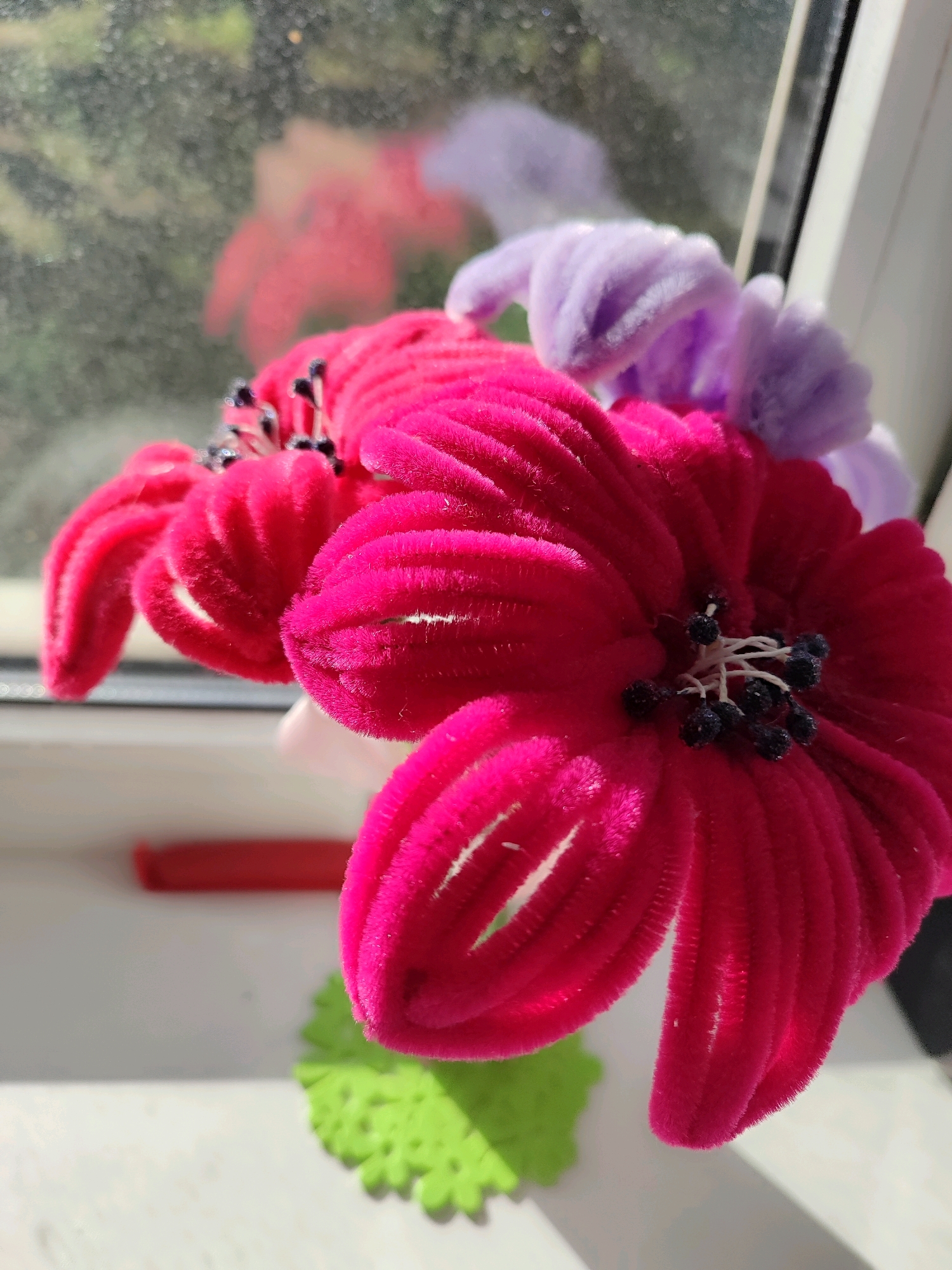 A close-up, eye-level, outdoor shot shows several artificial flowers with bright pink and lavender petals arranged near a window. The flowers are made of fuzzy pipe cleaners and have dark bead stamens in the center. Sunlight streams in from the left, casting shadows and creating highlights on the petals. In the background, out of focus, is a blurred reflection of a red flower in the window. A green, flower-shaped decorative mat is visible on the windowsill, along with a red cylindrical object. The scene appears to be in a domestic setting, possibly a windowsill in a home.