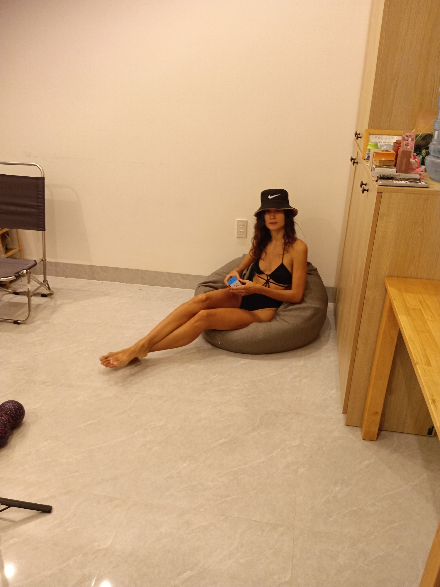 The image captures a young adult woman relaxing indoors in Mũi Né, Vietnam. She is seated on a grey beanbag chair, wearing a black two-piece swimsuit with criss-cross straps and a black bucket hat prominently displaying a white Nike swoosh logo. Her long, dark wavy hair is visible, and her bare legs are extended forward on the tiled floor, crossed at the ankles. She holds a small, light blue and white object in her hands.
The setting is a room with light cream-colored walls and a light grey tiled floor featuring subtle marbling patterns, with a darker grey tile trim along the wall base. To the left, a dark grey fabric and silver metal folding chair is partially visible. On the right, a light wooden cabinet with dark handles holds various items on top, including a large clear water bottle, a framed object, and several small boxes or remotes. A light wooden table or bench extends from the cabinet into the foreground. A white electrical outlet is mounted on the wall behind the woman. In the bottom-left corner, a dark purple and black patterned cylindrical object, possibly a foam roller, rests on the floor. The artificial lighting suggests an indoor scene, and the woman appears relaxed.