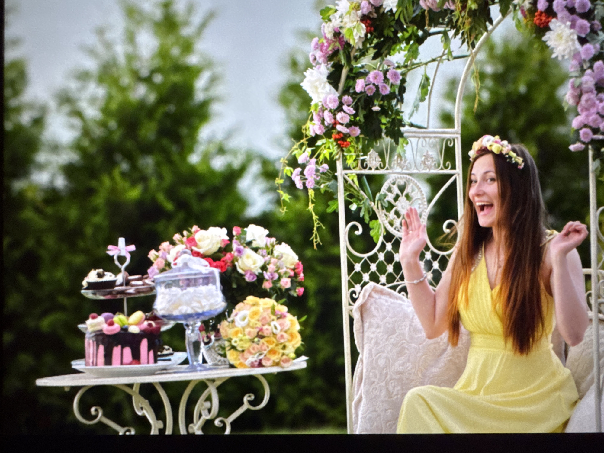 In an outdoor garden setting, a young woman wearing a yellow dress and a floral crown smiles with her mouth open, her right hand raised. She is seated on a white, ornate bench. In front of her, on a white table, is a dessert spread including a cake, cupcakes, and a glass jar filled with whipped cream. Behind the table, a floral archway is adorned with pink and white flowers. The background consists of blurred green trees. The scene does not contain any elements that place it specifically in the city of Riga, Latvia.