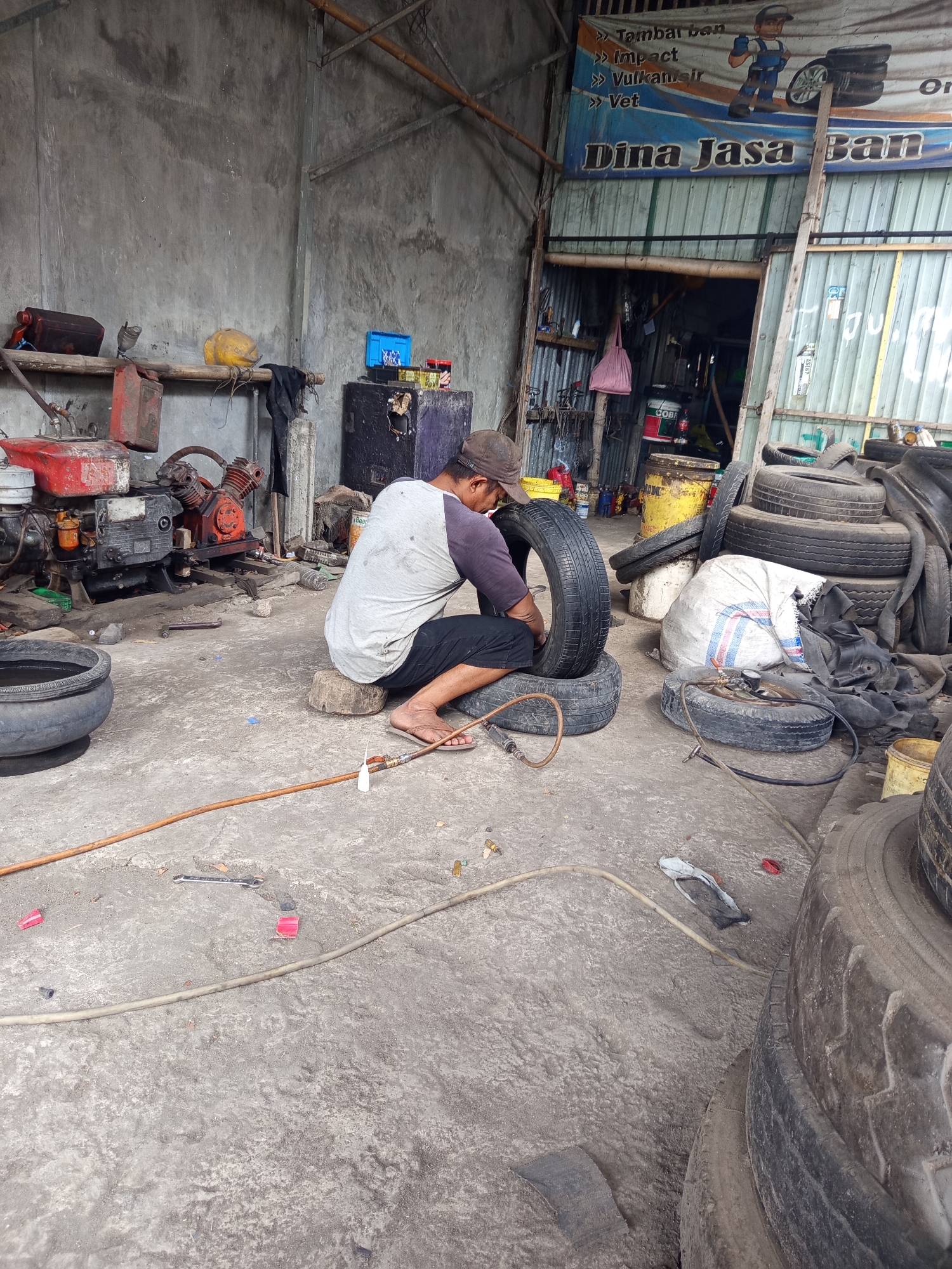 A man is working on a tire in a tire repair shop located in Mrican, Indonesia. The shop appears to be a small, open-air facility with a concrete floor and a corrugated metal roof. The walls are made of rough concrete, and there are various tools and equipment scattered around, including an air compressor, spare tires, and a large pot filled with water. The man, wearing a baseball cap, a grey t-shirt, and black shorts, is seated on a wooden block, using a tool to work on a tire that is placed on top of another tire. A hose with a pressure gauge is connected to the tire he is working on. In the background, a banner with text in Indonesian advertises tire repair services. The overall scene depicts a typical workshop environment for tire maintenance.