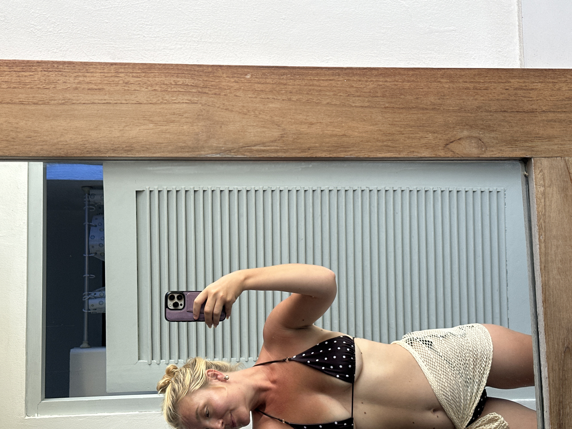 A woman with blonde hair tied in a bun takes a mirror selfie in a room. She is wearing a black and white polka dot bikini top and a light-colored netted sarong tied around her waist. Her left arm is raised, holding a purple-cased smartphone. The background consists of louvered doors and a wooden frame. The scene appears to be a domestic setting in North Kuta, Indonesia.