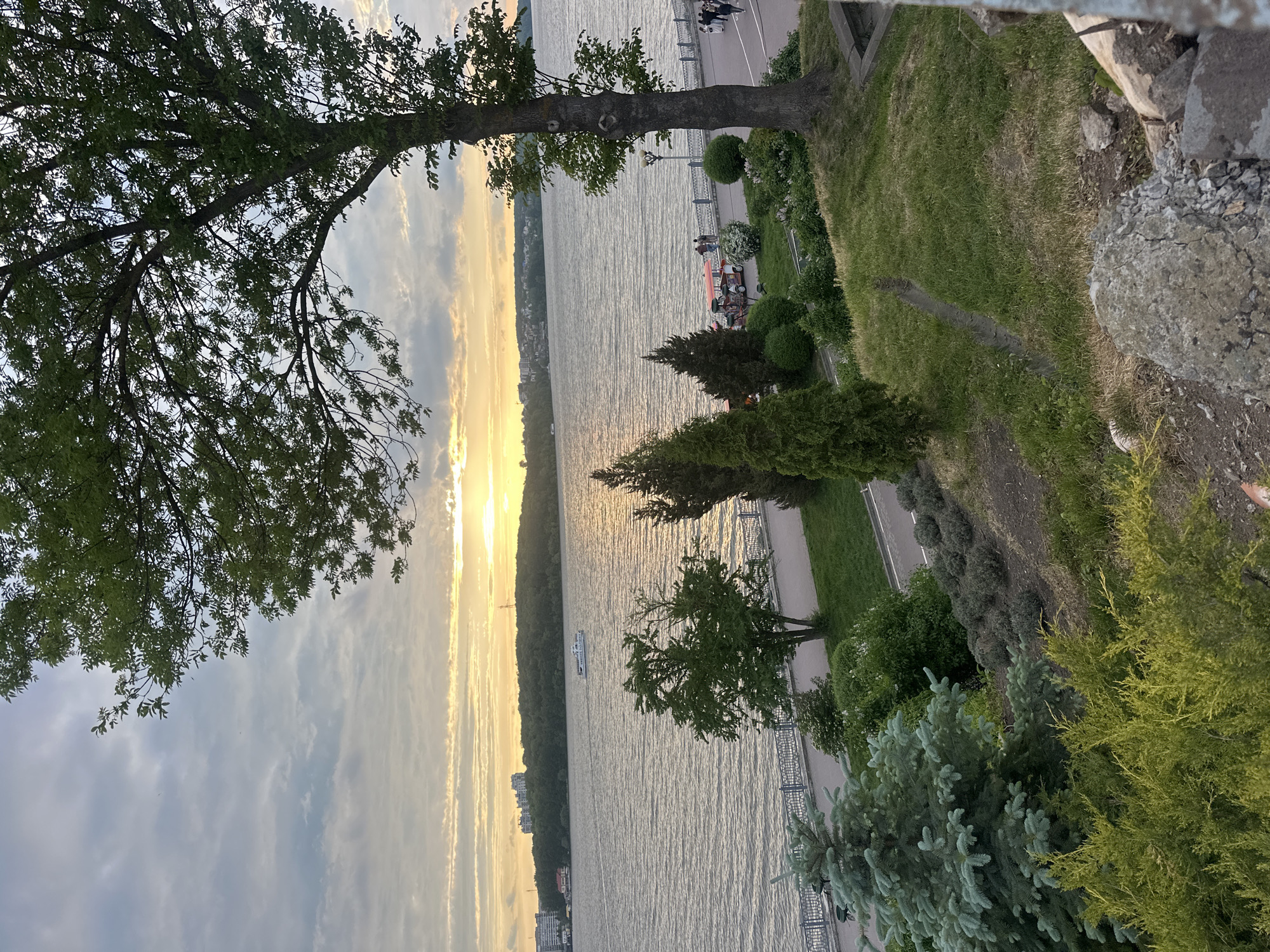 The image shows a sunset view of Ternopil Lake in Ternopil, Ukraine. The lake stretches across the center of the frame, with a tree branch and foliage on the left and the embankment on the right. The sky is filled with clouds and the sun is setting on the horizon. Along the embankment, there is a sidewalk with people strolling and benches for seating. A boat is visible on the lake. The embankment is adorned with a grassy area, various trees, and decorative shrubs. A stone wall borders the edge of the embankment in the foreground.