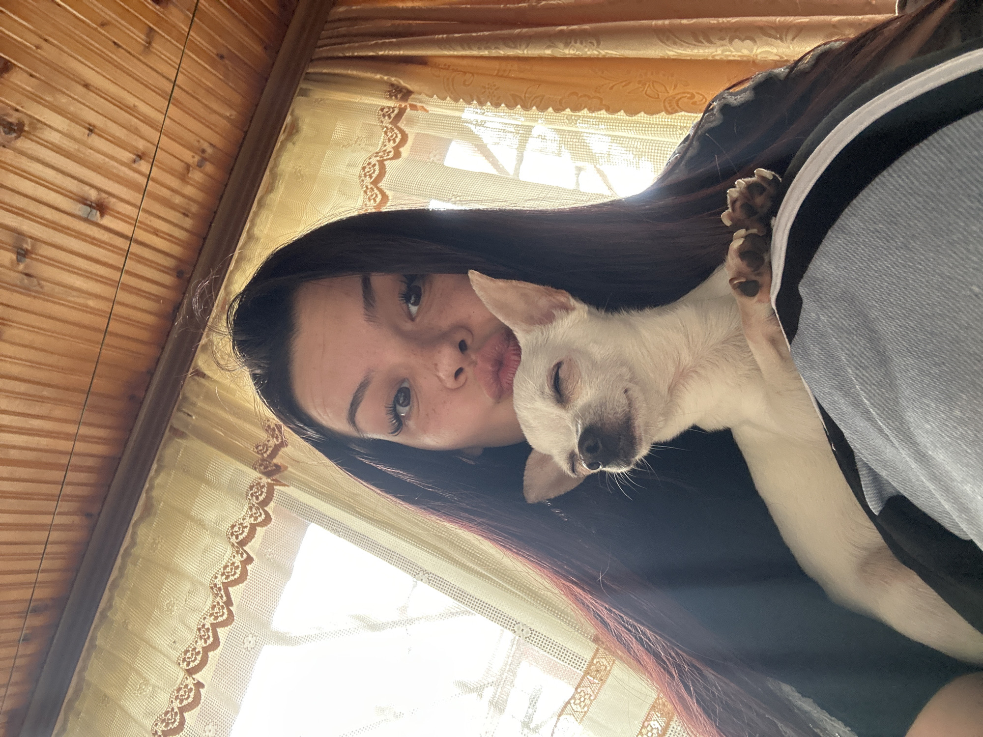 A rotated, close-up selfie shows a young woman with long, dark hair and a small, white dog. The woman is pouting her lips towards the camera, while the dog is nestled close to her face, its eyes partially closed. The dog's paw is visible against the woman's dark clothing. The background features a wooden ceiling, a window with patterned curtains, and a partially visible piece of light blue and black clothing on the right side of the image. There is nothing in the image to indicate that this photo was taken in Skala, Bulgaria.