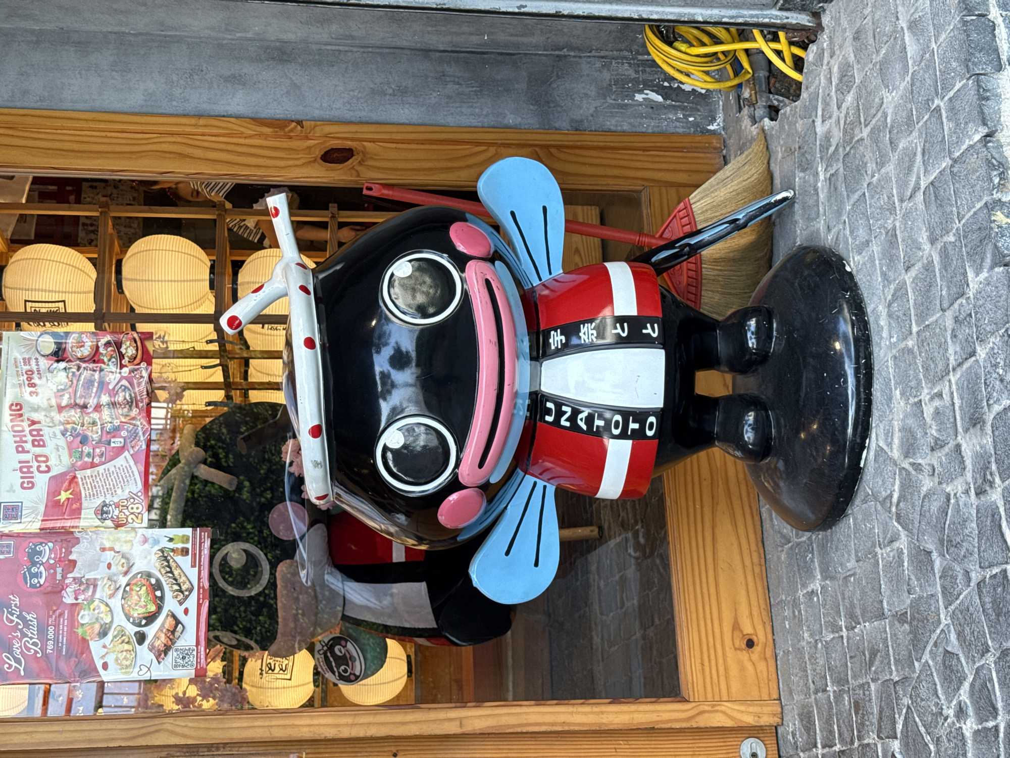 The image shows a colorful, stylized statue of a Japanese-themed figure, likely a restaurant mascot, standing in front of a building. The statue has a black head, large white eyes, pink cheeks and a pink rectangular mouth. It wears a traditional Japanese headband with red polka dots and light blue "wings" on the side of its head. Its body is decorated in red, white, and black stripes with Japanese writing; the word "UNATOTO" is also prominently displayed in English. A broom leans behind the statue. In the background, there is a wooden-framed window filled with white spherical lanterns, advertisements, and foliage. The location appears to be in the city of Thủ Đức, Vietnam based on the promotional materials visible in the background.