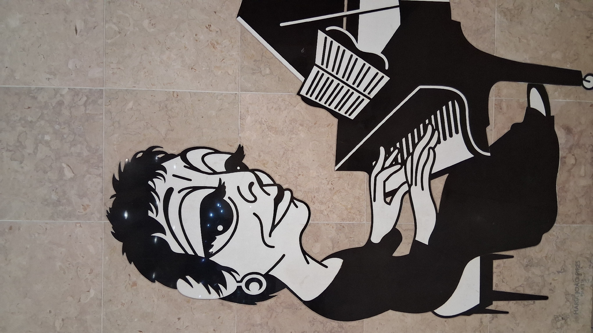 An artistic black and white graphic illustration of a woman playing a grand piano is affixed to a beige tiled wall in Lisbon, Portugal. The woman has a large head with wide eyes that have small blue sparkling lights embedded in them. She has short, spiky black hair, a prominent nose, and a wide smile. She wears a black dress and a large hoop earring in her left ear. Her hands are positioned on the piano keys. A sheet of music is open on the stand in front of her. The piano is black and white, with white keys. The artwork is signed "MARIA JOAO PIRES" at the bottom left.