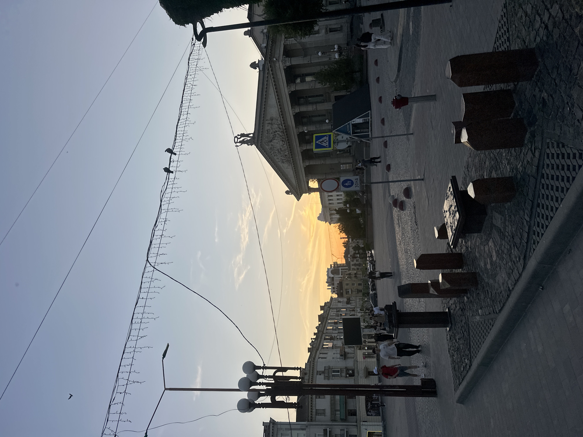 The image depicts a public square in Ternopil, Ukraine, during sunset. In the foreground, there is an outdoor chess set with carved wooden pieces and a stone table. Several people are visible walking around the square. Behind the chess set, a tall lamppost with multiple lights stands. In the background, there are several buildings with classical architecture, including a prominent building with columns and a pediment, likely a cultural institution. The sky shows the warm hues of sunset, with some clouds. Overhead wires and decorative lights are strung across the square. The ground is paved with cobblestones and patterned tiles. A traffic sign indicating a pedestrian crossing is visible. Several red bollards are placed along the edge of the walkway.