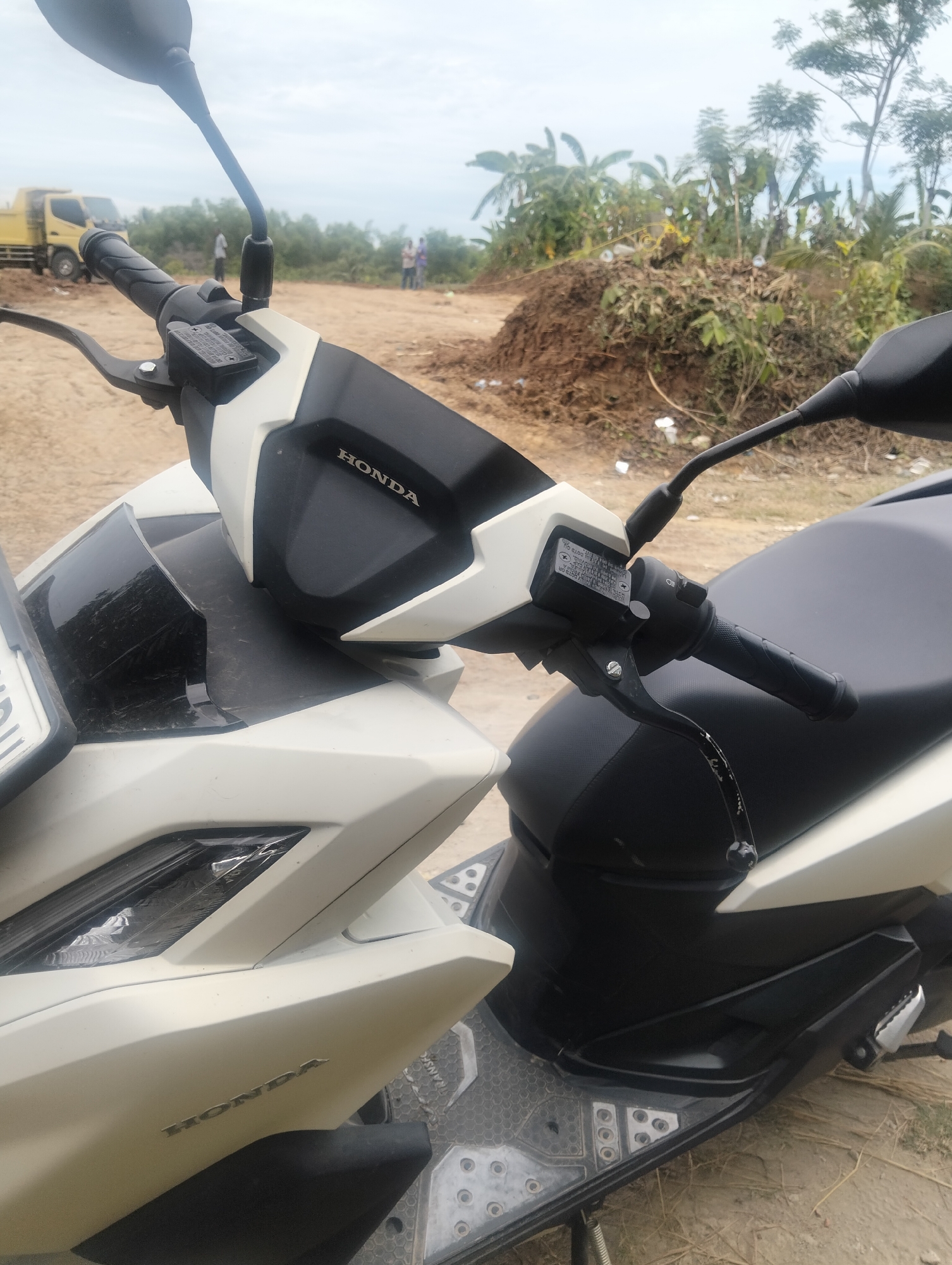 A close-up, eye-level shot captures the front and right side of a white and black Honda scooter. The scooter is parked on a dirt path. The handlebar with the Honda logo is visible, along with the brake lever and a reservoir. The seat of the scooter is black. In the background, a yellow truck is partially visible, along with some vegetation and trees. The sky is overcast. The location is identified as Payagaboh, Indonesia, an area likely undergoing construction or development given the presence of the truck and piled earth.