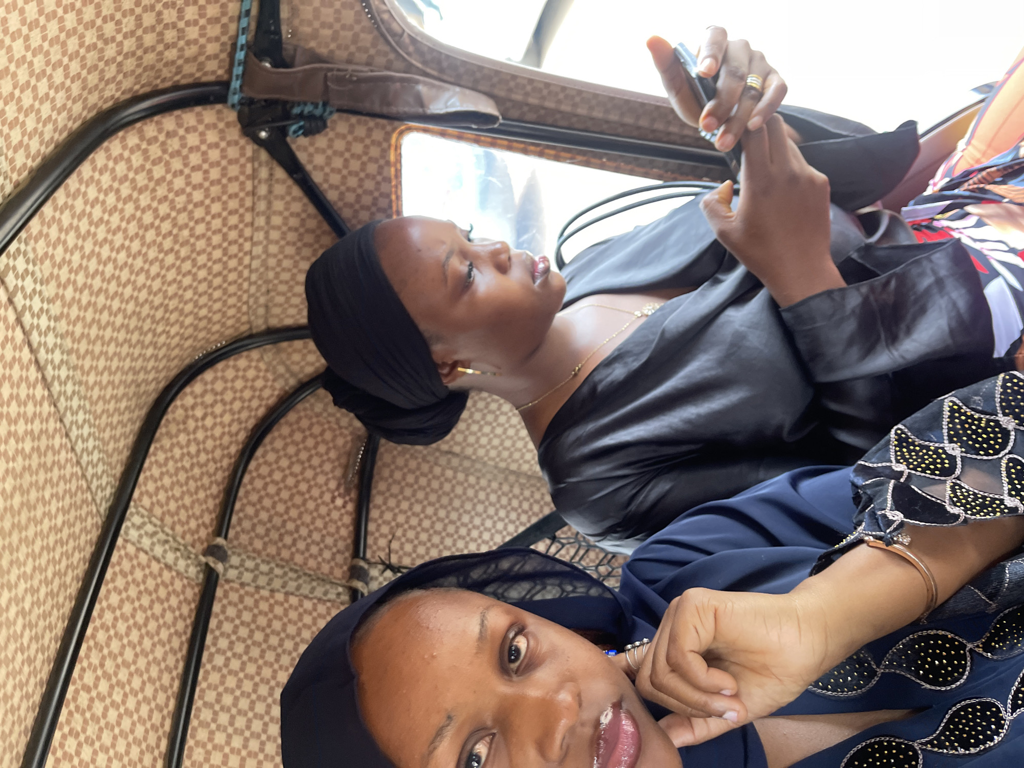Two women are inside a tuk-tuk, likely in Rafin Par, Nigeria. The woman on the left is facing the camera and is wearing a dark blue headscarf and a dark blue dress with gold and black embellishments. She has her hand near her face, touching her chin. The woman on the right is looking down at her phone and is wearing a black headscarf and a black satin top. She is holding a black mobile phone in her hands. The interior of the tuk-tuk has a patterned beige fabric lining the roof and sides. The overall lighting suggests it is daytime.