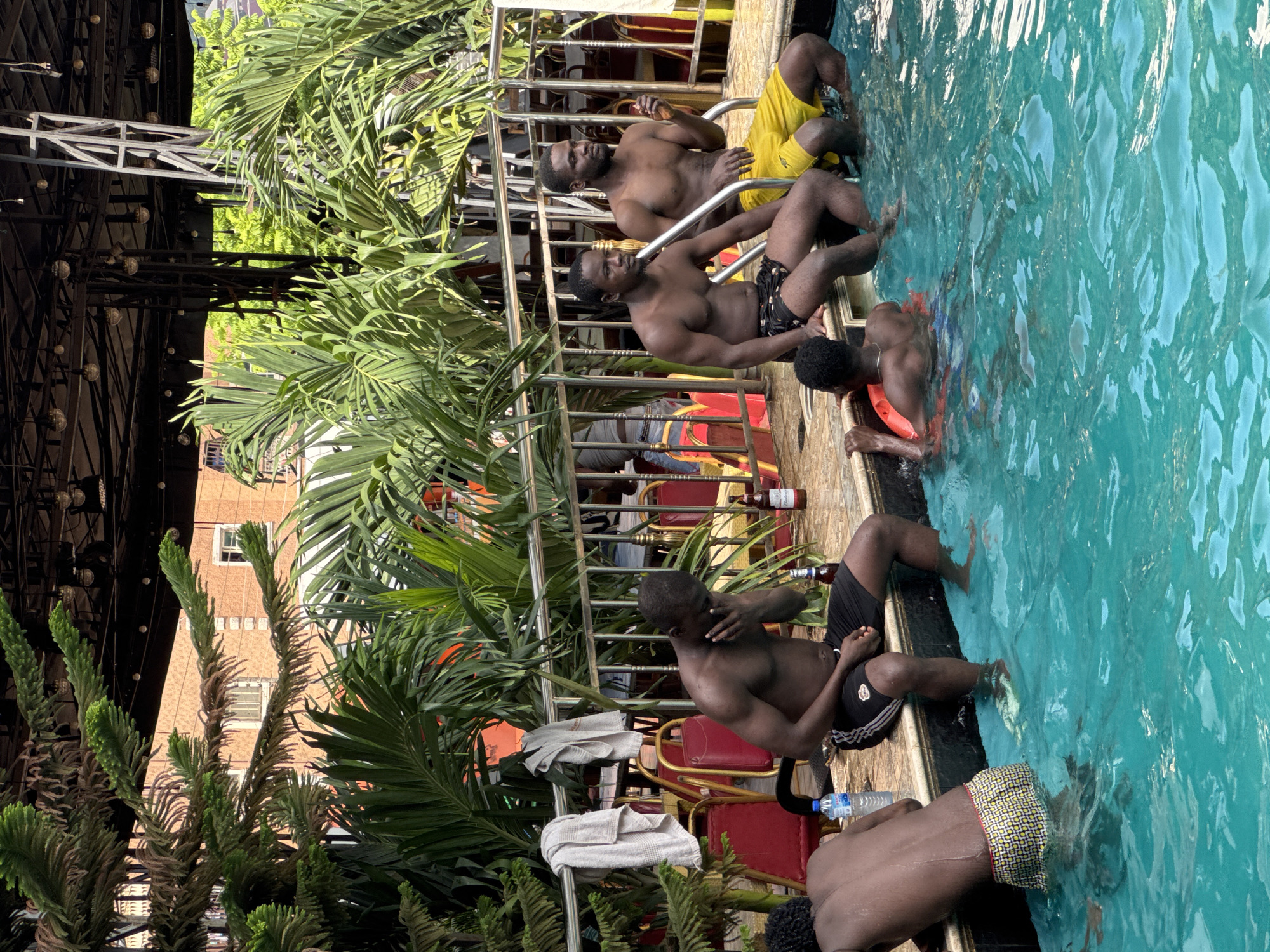 In Nkpor, Nigeria, at a poolside establishment, several young men are gathered. The image captures a group of individuals, predominantly young Black men, interacting in and around a swimming pool. One person is partially submerged in the turquoise water, facing away from the camera and wearing patterned swim trunks. To his right, two men are seated on the pool's edge, with their legs in the water. The man on the left wears black swim shorts with white stripes, while the man on the right is in yellow swim shorts. Both appear to be engaged in conversation or observation. Another man is seated further to the left, closer to the pool's edge, wearing black swim shorts and appears to be touching his face. On the pool deck behind the seated individuals, two bottles of beer and a bottle of water are visible, along with red chairs and metallic railings. Lush green palm fronds and a pine tree frame the scene, suggesting an outdoor setting with a tropical or subtropical ambiance. The background reveals a building with orange-toned walls and windows, and a dark, overhead structure with lighting fixtures. The overall atmosphere suggests a relaxed, social gathering.