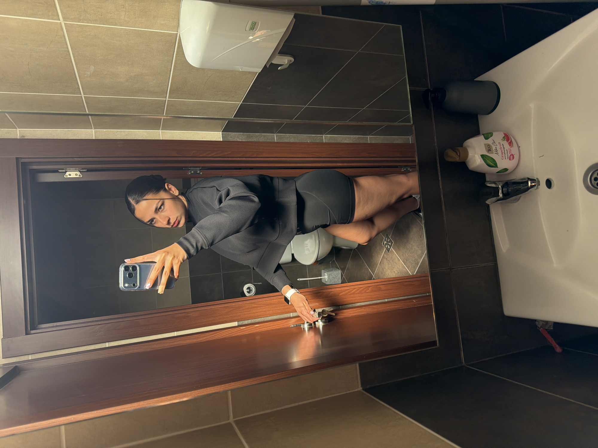 A woman in black athletic attire is taking a selfie in a bathroom mirror. The bathroom has brown and dark grey tile. A white soap dispenser and sink with toiletries are visible to the right of the mirror. A toilet is located to the right and further back. The location cannot be determined to be the city of Gabrovo, Bulgaria from the visual information provided.