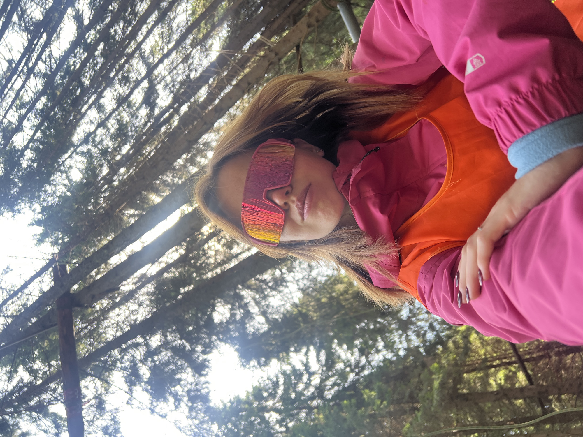 The image shows a person wearing a pink jacket, an orange vest, and futuristic ski goggles with a pink and orange lens. The person's hair is blonde. The background is out of focus, but appears to be a forest with tall trees and dappled sunlight. The image is taken from a low angle, looking up through the trees. The lighting suggests it is daytime. The person's face is partially visible, and they appear to be looking towards the camera. There is no visible text in the image. The overall impression is of someone enjoying an outdoor activity in a natural setting.