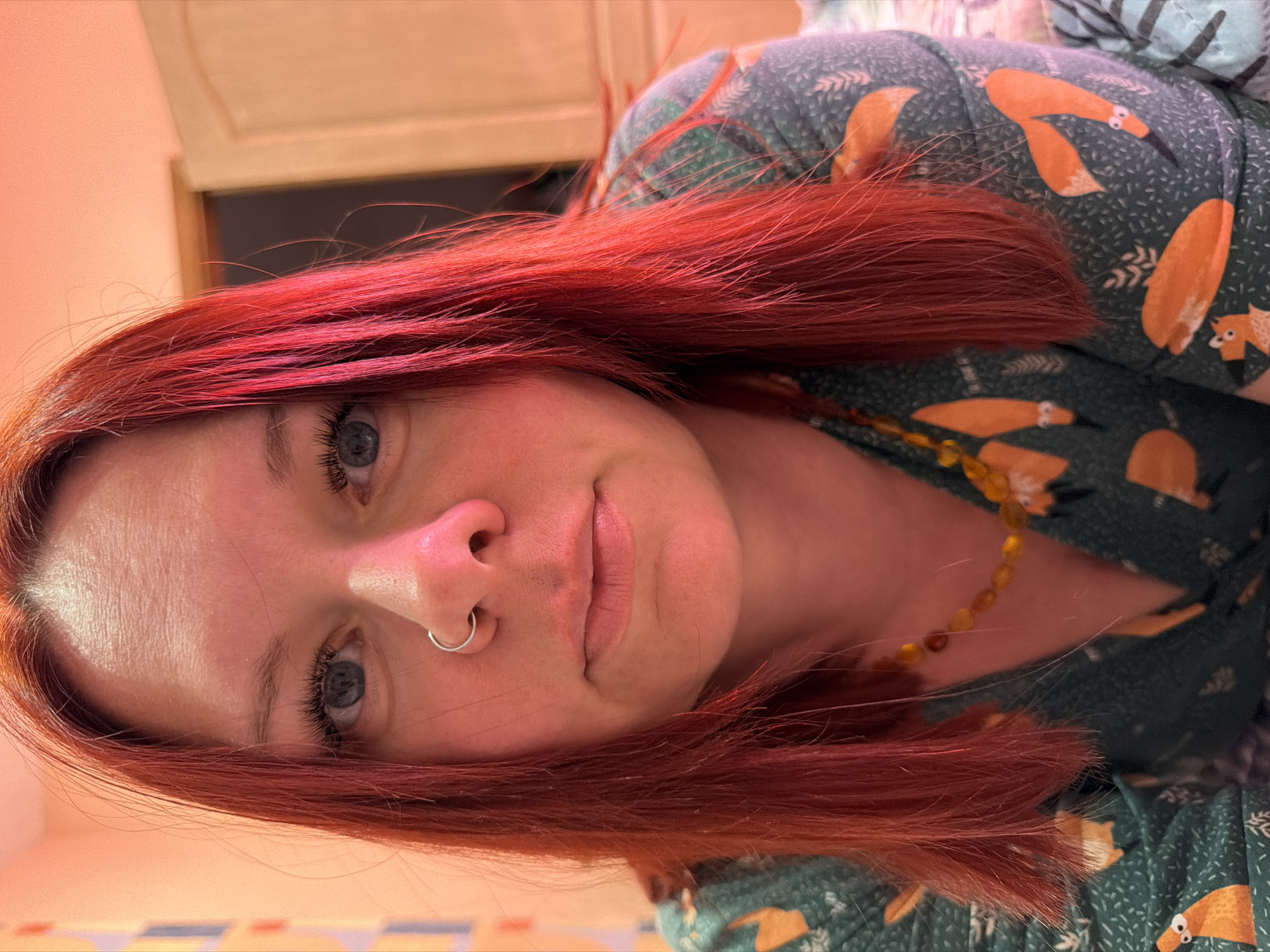 A person with fair skin, red hair, and blue eyes is centered in the image. They are wearing a silver nose ring and a beaded necklace. The person is wearing a green shirt with an orange fox pattern. The background includes a light beige wall and what appears to be a pillow with a floral design. The location is not evident in the image.