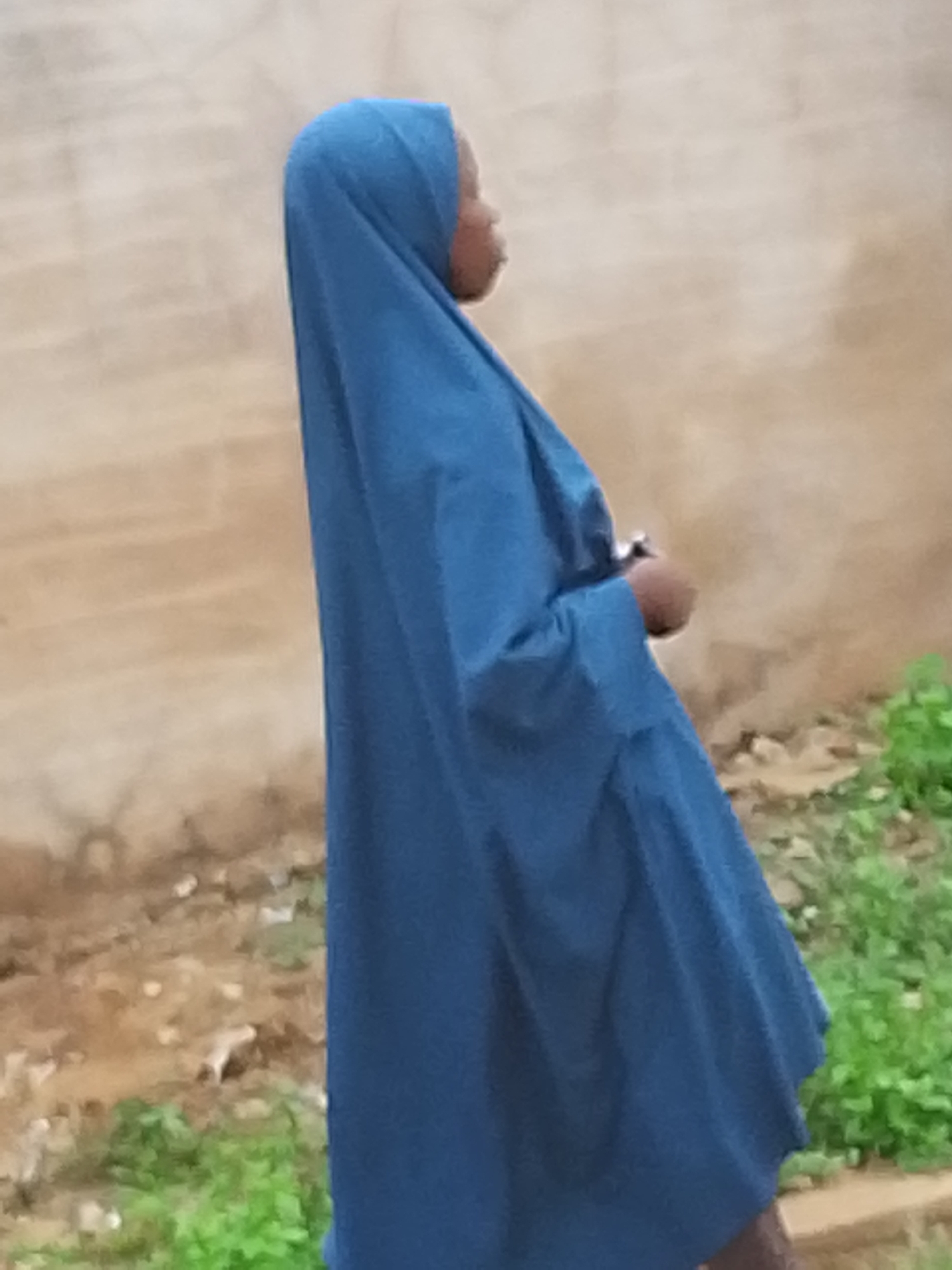 A person wearing a blue hijab stands in front of a light brown wall, possibly in an outdoor location in Jos, Nigeria. The person is facing left. Green vegetation grows along the ground in front of the wall.