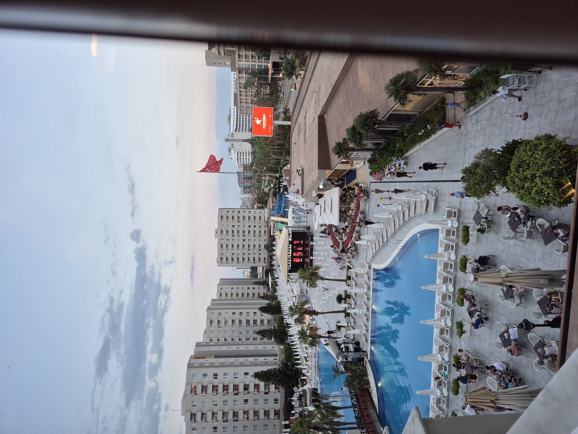 The image shows an aerial view of a resort pool area with people dining at tables and lounging by the pool. The pool is large and brightly lit with blue water, reflecting palm trees. Around the pool, white lounge chairs and umbrellas are set up.
In the background, several tall, modern buildings are visible, suggesting an urban or coastal resort setting. A large red billboard with white text and a star logo is prominent in the mid-ground. A Turkish flag is flying on a flagpole in the foreground.
People are seen sitting at outdoor tables, some eating and drinking, while others are walking around the resort. There are palm trees and green bushes scattered throughout the area, adding to the tropical atmosphere. A stage with lighting is set up near the pool.
The sky is overcast with soft clouds, and the lighting suggests it might be late afternoon or early evening. The overall mood appears relaxed and social, indicative of a holiday resort.
Visible text on the billboard reads "Corendon".
