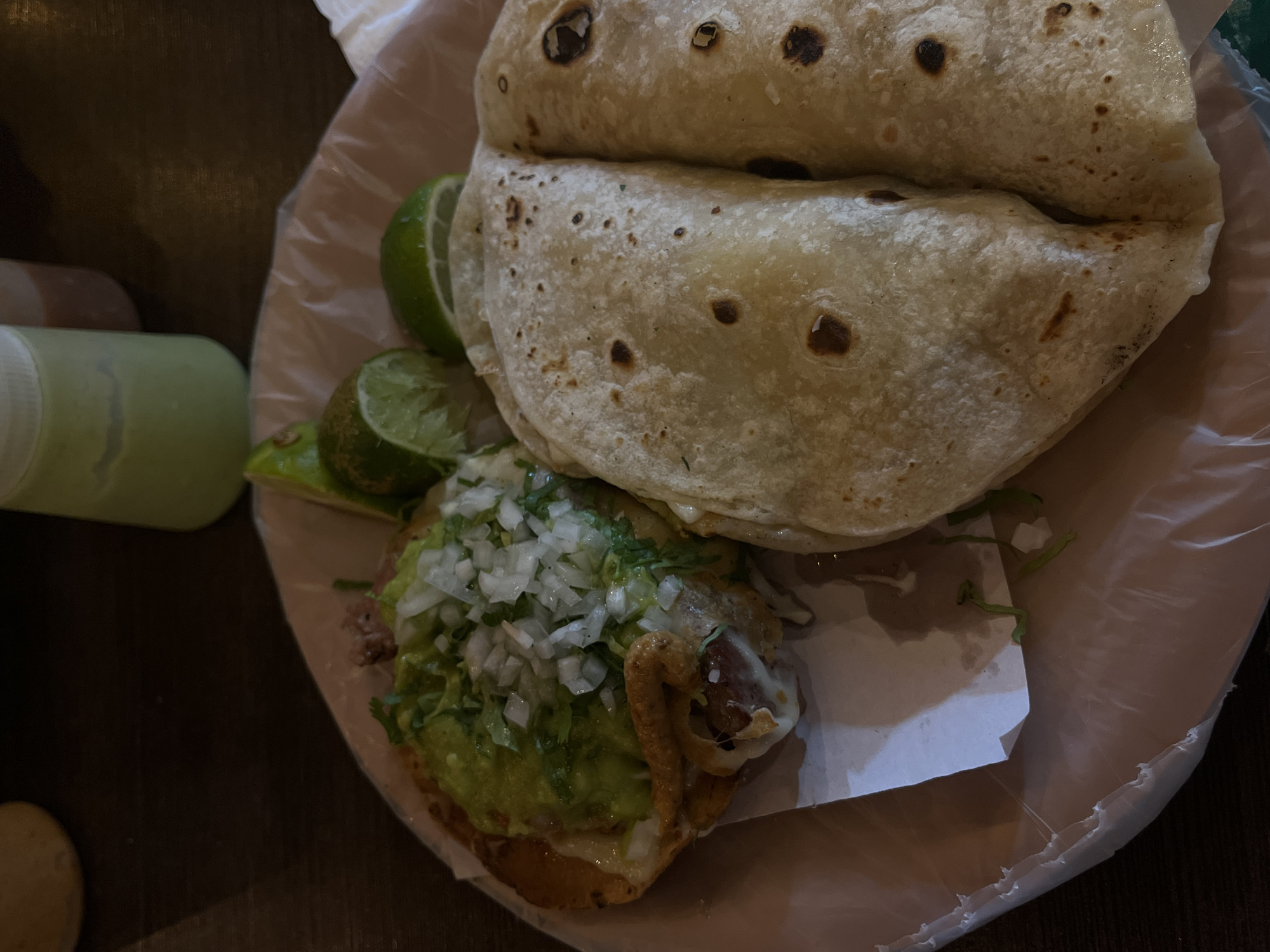 A close-up overhead shot shows a plate of Mexican food. Two tortillas, folded over one another, take up the upper right portion of the frame. To the left, a taco with a topping of guacamole and diced white onions is visible. Two lime wedges are positioned between the taco and the tortillas. To the left of the plate, a green bottle sits on a dark surface. The food is served on a disposable plate lined with clear plastic. The setting might be in San Bernardino Tlaxcalancingo, Mexico.