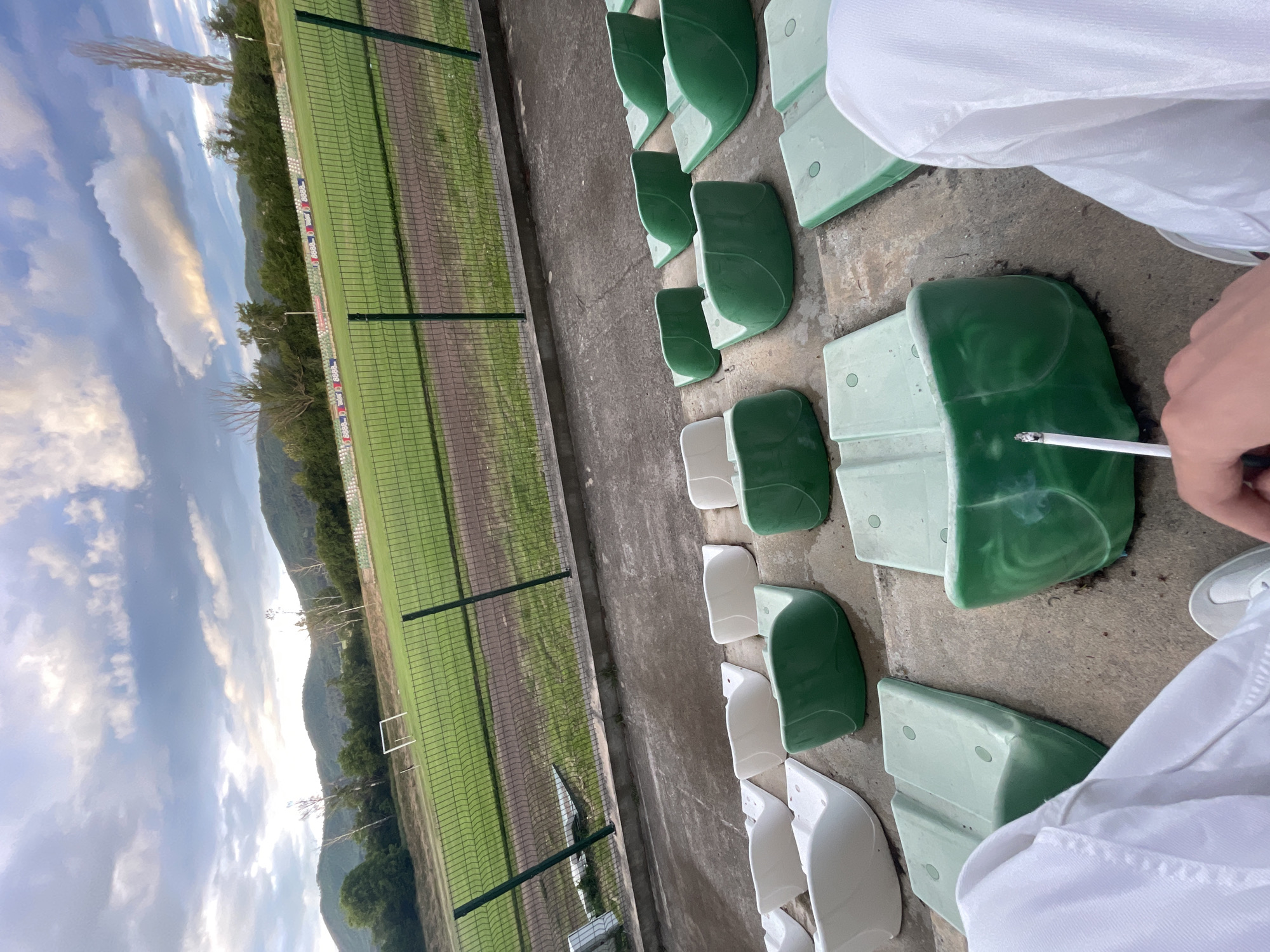 The image, rotated 90 degrees clockwise, presents a view from a stadium grandstand towards a sports field with a natural landscape background. In the immediate foreground on the right, a person is partially visible, clad in a white long-sleeved garment. Their right hand holds a lit cigarette, from which a plume of smoke rises gently.
The foreground and mid-ground are filled with rows of empty stadium seats, predominantly green but also featuring some white ones, all made of plastic and mounted on concrete steps. Beyond the seating area, a tall green metal mesh fence separates the spectators from a green grass sports field, likely a football (soccer) pitch, which shows faint markings. A white goal frame is visible on the far side of the field. Along the sideline of the field, behind the fence, are small, faded, colorful rectangular elements that appear to be banners or advertisements, though no specific text is discernible.
The background is dominated by lush green mountains or hills stretching across the horizon, suggesting a natural and possibly rural setting. Above the mountains, the sky is partly cloudy with soft, diffused light, indicating either late afternoon or early evening. The field appears empty, and there is no visible sporting activity. The scene conveys a quiet, perhaps contemplative, moment. The mountainous scenery is consistent with the provided location context of Ihtiman, Bulgaria.