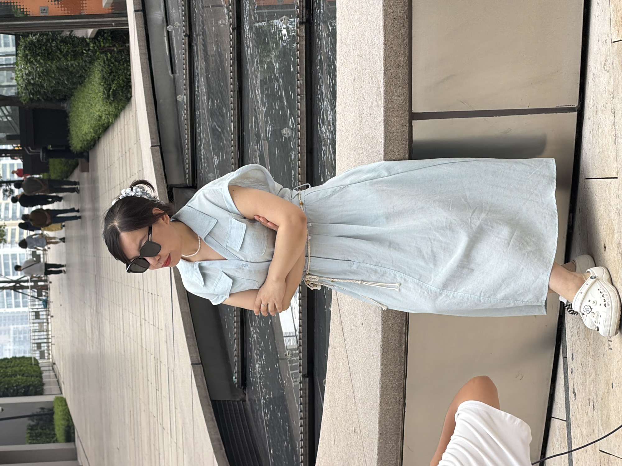 A woman with dark hair pulled up in a bun and adorned with a silver clip, wearing large dark sunglasses, a light blue collared dress, a pearl necklace, and white Crocs, is sitting on a textured stone ledge. Her arms are crossed, and she is looking off to her left. A person in a white t-shirt is partially visible on the left side of the frame, crouching down. The background features a paved walkway with several individuals walking in the distance, manicured green hedges, and modern buildings. The scene appears to be outdoors, possibly in a public plaza or urban park in the Yuzhong District, China, indicated by the architectural style and foliage.