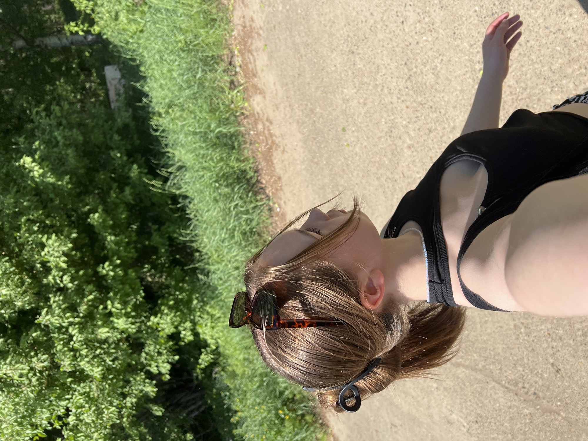 A woman in a black tank top is taking a selfie outdoors on a concrete path. Her hair is light brown and pulled back in a ponytail with a black clip. She has sunglasses perched on her head. There is green grass and foliage to her left. The location cannot be determined definitively as Chaikovsky, Russia, as there is no contextual information in the image to confirm the location.