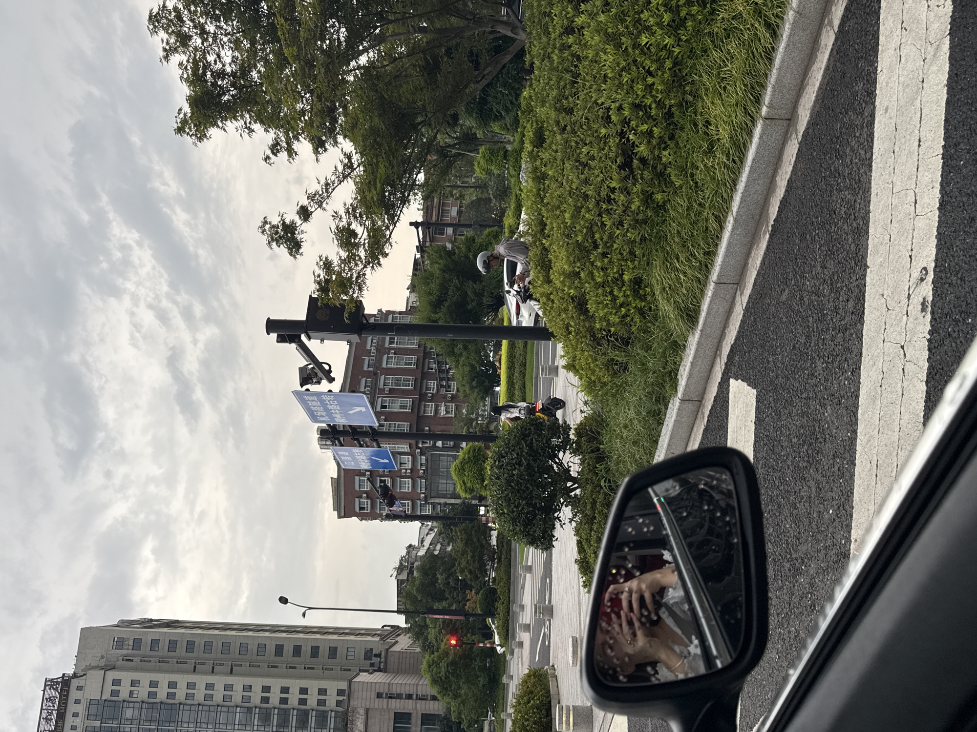 This image captures a view from inside a vehicle looking out at an intersection in Yiwu, China. The sky is overcast with a high density of clouds. In the foreground, the driver's side mirror reflects a person holding a smartphone, seemingly taking a selfie, with their hands and part of their face visible. The mirror also shows streaks of rain or water droplets.
The road features white pedestrian crossing lines and a grey curb. To the right of the road is a meticulously maintained green hedge. In the background, several buildings are visible. A tall, modern high-rise with numerous windows stands on the left, with a sign at the top that reads "HUI HOTEL". Adjacent to it, a traffic light shows a red signal. Further back, a red brick building with many windows is situated, and above it, traffic signs with blue backgrounds and white text are mounted on poles.
On the street, a woman wearing a white top and black bottoms is riding a yellow and black electric scooter. Further to the right, a person wearing a grey hooded sweatshirt and a helmet is on a scooter, looking down at their phone. Another white car is parked or stopped behind them. Trees with dense green foliage are scattered throughout the scene, contributing to the urban greenery. The overall impression is an ordinary street scene in a city during cloudy weather.
