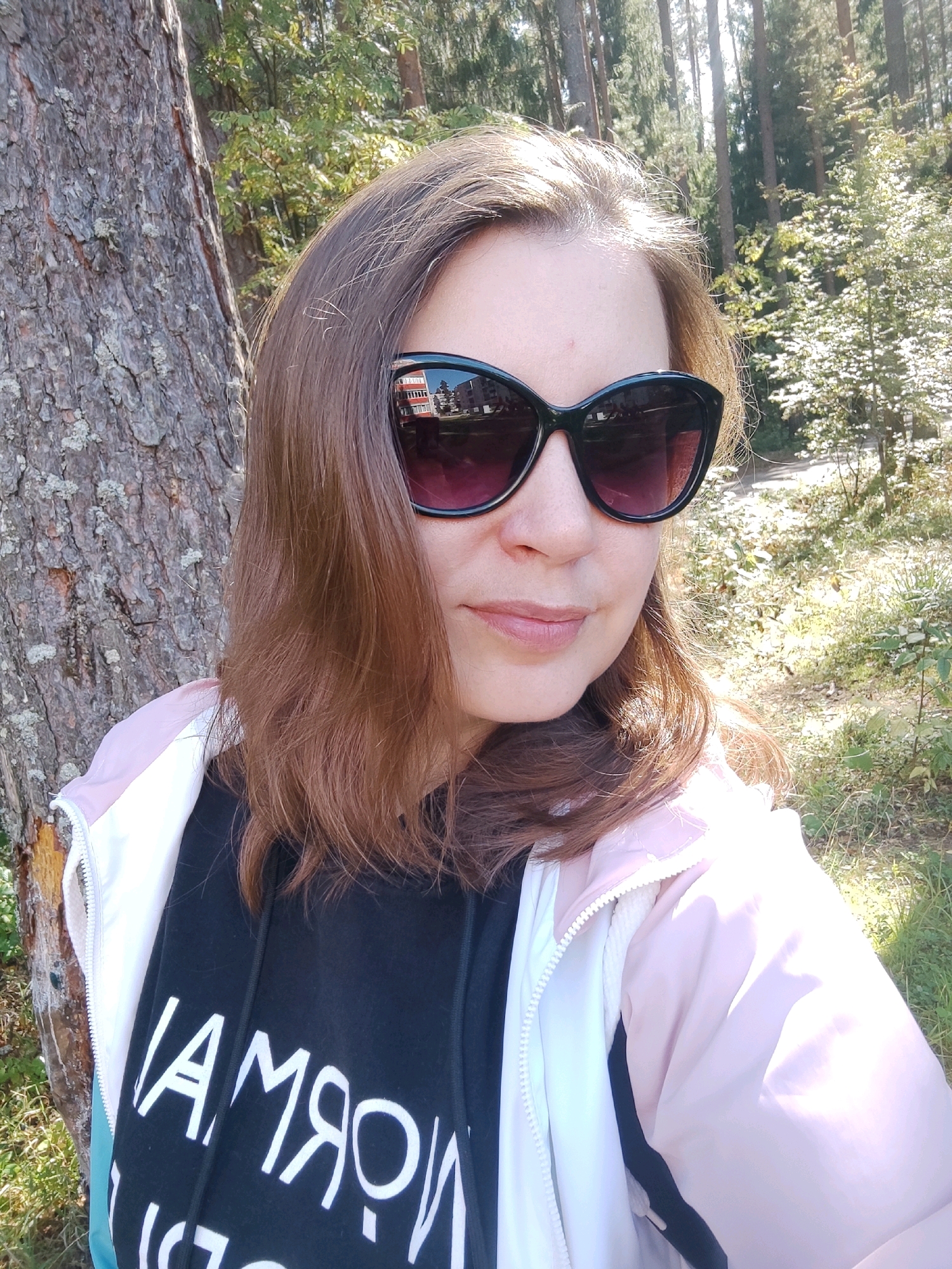 A woman is taking a selfie outdoors in a wooded area. She has brown hair and is wearing sunglasses and a pink and white jacket over a black hoodie with white text. The background shows trees and foliage.