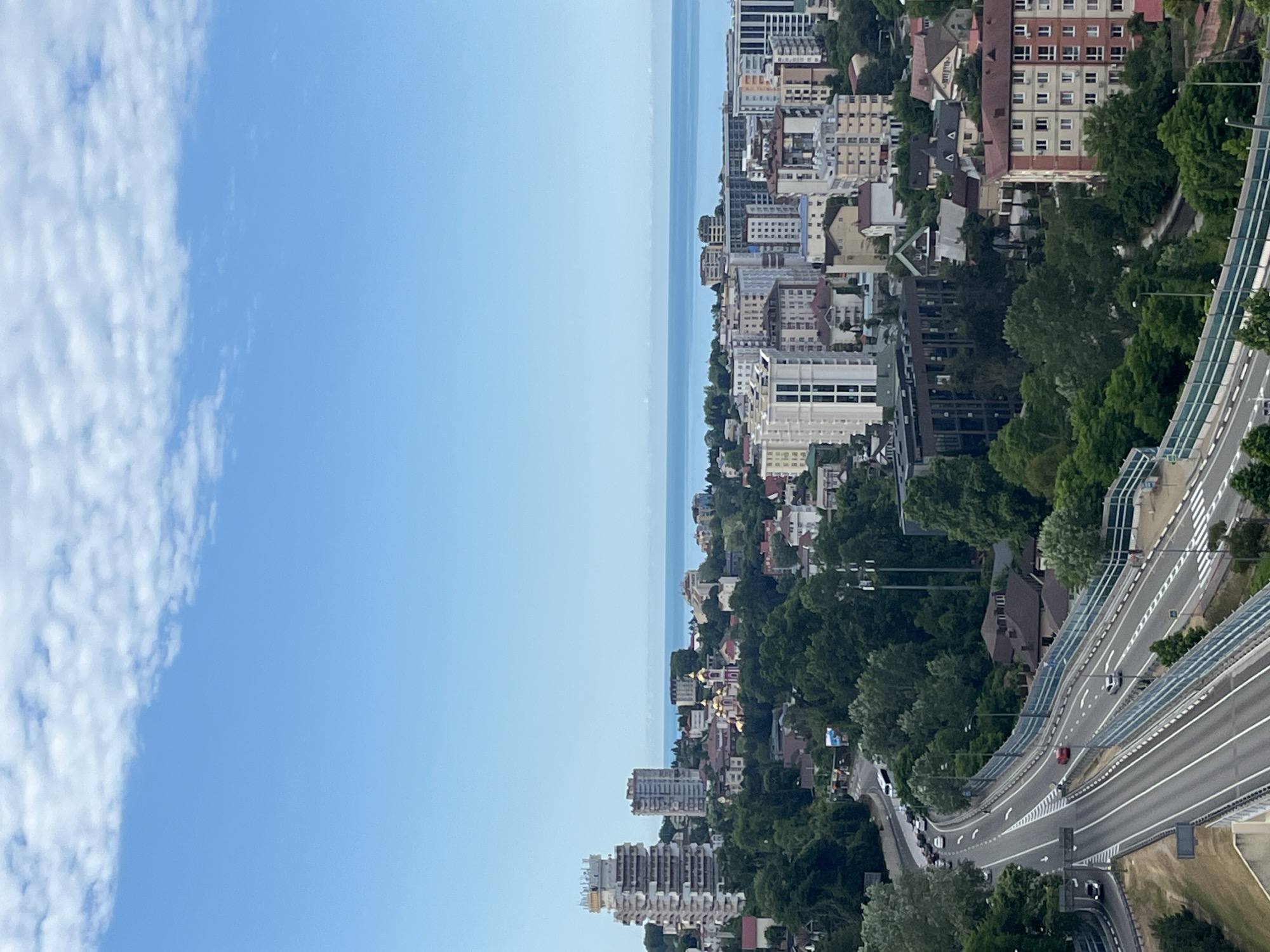 The image shows a panoramic view of a coastal city with a highway in the foreground. The city, identified as Sochi, Russia, features a mix of modern high-rise buildings, older residential structures, and a prominent Orthodox church with golden domes. Lush green trees are interspersed throughout the urban landscape, leading down to the blue sea in the background. The sky is a clear blue with scattered white clouds. The highway is multi-lane and appears to be elevated in some sections, with vehicles visible on it. The overall scene depicts a sunny day in a vibrant coastal city.