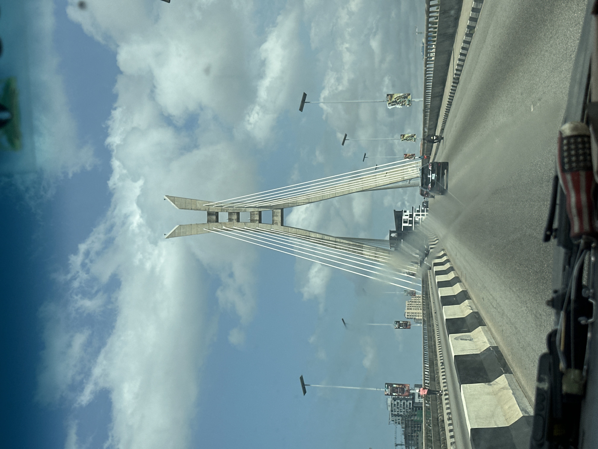 A slightly skewed, first-person view of a bridge, with a blue sky with clouds in the background and a section of the Ikoyi-Lekki Link Bridge visible. A car traverses the bridge, which shows other cars on the same trajectory. Street lights and billboards also line the bridge. A small American flag is visible on the vehicle's dashboard.