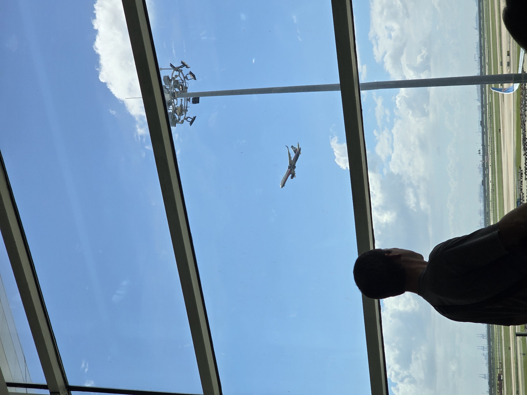 An aerial view from inside an airport terminal captures a man in silhouette looking out a large window at an airplane taking off. The man is positioned in the foreground, facing away from the camera and towards the expansive sky visible through the window. The airplane, a commercial jet with a white body and dark tail fin, is seen ascending into the bright blue sky dotted with fluffy white clouds. In the background, an airport runway stretches into the distance, bordered by green fields and industrial structures. A tall light pole with multiple lights is visible to the right of the airplane, also against the sky. The scene suggests a moment of departure or arrival, with the man observing the powerful ascent of the aircraft against a backdrop of a typical airport environment.