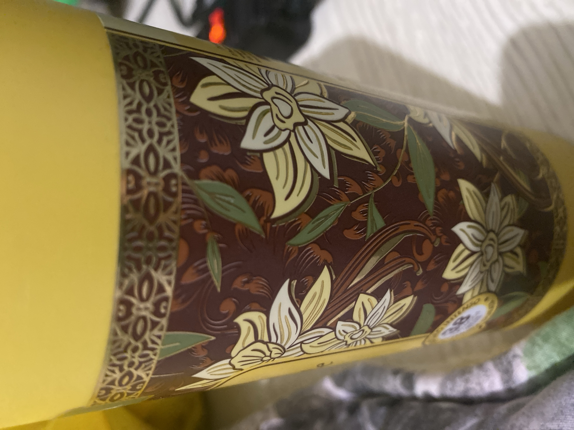 A cylindrical object, likely a tin can or canister, with a yellow body and decorative floral patterns is displayed. The floral design is composed of brown and cream-colored hues with green leaves. A band with a repeating geometric pattern borders the edge of the decorative design. The object rests on a textured surface, possibly fabric. The location and the city of Poltava, Ukraine, is not evident from the image.