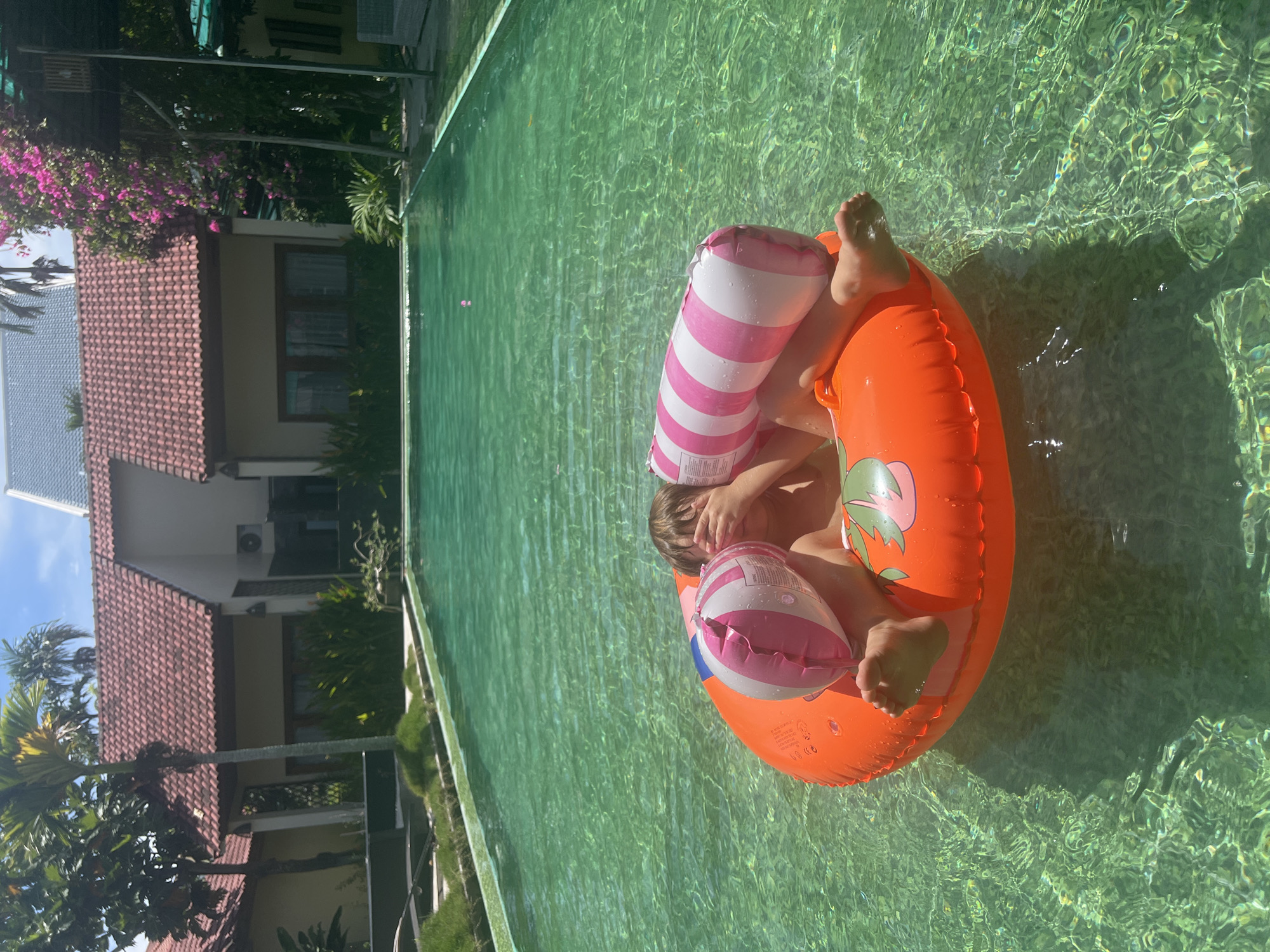 A child sits in an orange inner tube in a swimming pool. The child is partially obscured by their hands, which cover their face. Two inflatable pillows with pink and white stripes are placed around the child. The pool water is clear and green. In the background, there are buildings with red tile roofs, palm trees, and lush green vegetation. The scene appears to be in a resort or hotel in Hoi An, Vietnam, given the architectural style and tropical foliage. The lighting suggests it is a sunny day.