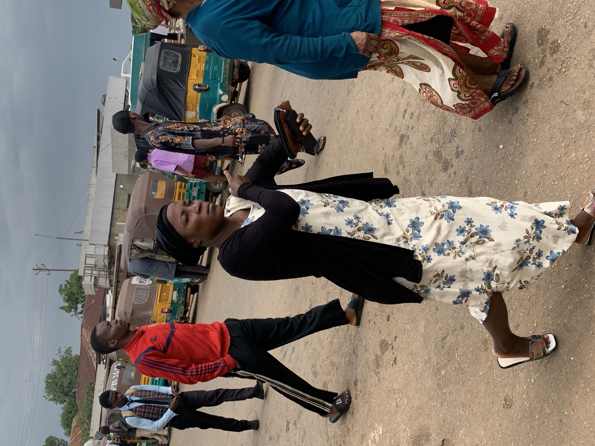 The image depicts a street scene in Gura Top, Nigeria, under an overcast sky. In the foreground, a woman with dark skin wears a black headscarf, a black cardigan over a light-colored dress with a blue floral pattern, and slides on her feet. She is walking and holding a brown wallet and a mobile phone in her left hand, with her right arm extended and finger pointing. To her left, a man in a red tracksuit with dark stripes on the sleeves and pants walks with his hands in his pockets. He wears black slides. Behind him, another young man in a light blue shirt, plaid vest, black tie, and black pants also walks, looking down at his hands. Further behind, other individuals are visible, including a person in a black and white patterned shirt and a person in a pink top. Several auto-rickshaws, common modes of transportation, are parked or moving in the background, some with their hoods up. The ground is paved with asphalt, and buildings with corrugated metal roofs are present in the background, along with power lines and poles. The overall atmosphere suggests a busy public area.