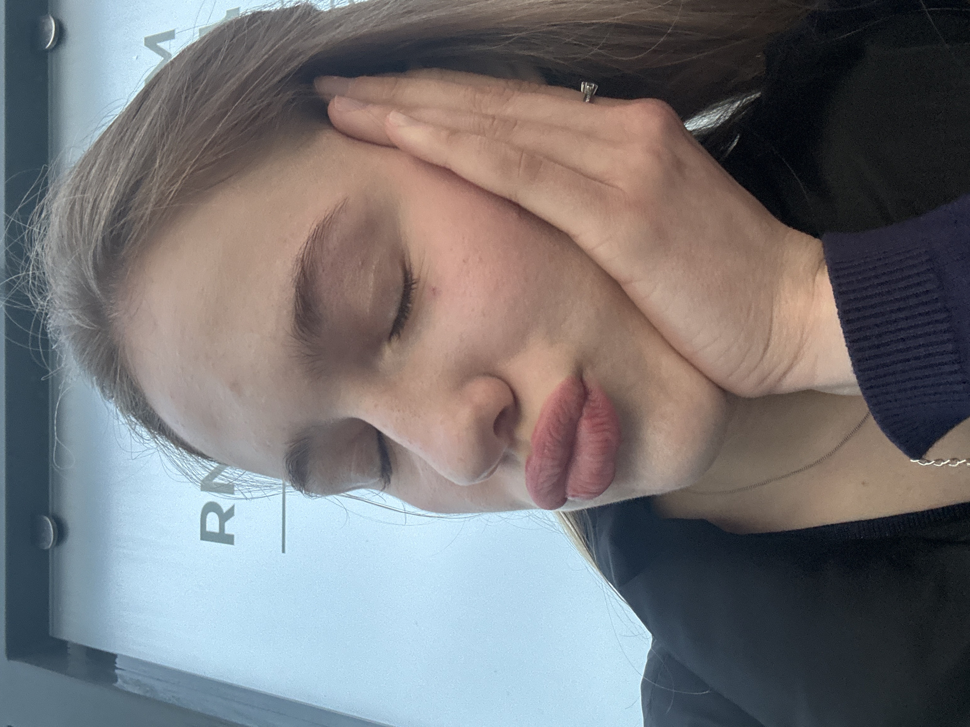 A close-up, low-angle, selfie-style shot captures the face of a young Caucasian female with blonde hair. Her eyes are closed, and her right hand is pressed against her cheek, with her thumb resting near her ear and fingers extended down her jawline. Her lips are pursed, and she appears to be wearing lipstick. She has light skin with some freckles and a small red mark on her left cheek. She is wearing a black jacket over a dark blue garment with a silver necklace. The background appears to be a light blue surface with the letters "RN" and a horizontal line visible, suggesting it might be part of a sign or a wall in an urban environment. There are no discernible elements that place the scene specifically in the city of Arfa, Russia.