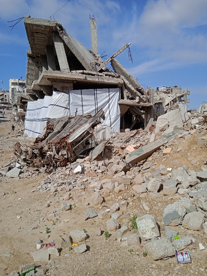 The image portrays a scene of extensive urban devastation in Khan Yunis, Palestinian Territory, under a clear blue sky with scattered white clouds. The foreground consists of uneven, light-colored earth intermixed with a significant volume of concrete rubble, broken bricks, and fragmented construction materials. Sparse green vegetation and a few pieces of litter, including wrappers, are present on the ground.
A severely damaged multi-story reinforced concrete building occupies the midground. Its upper levels are extensively collapsed and twisted, with exposed rebar protruding from fractured concrete slabs and beams, indicating structural failure. The lower section of this building features several openings covered with white, corrugated tarpaulin-like sheets, some bearing faint Arabic text. Immediately in front of this structure, a heavily rusted and compacted vehicle chassis, appearing to be from a car or small truck, is embedded within a pile of debris.
In the background to the left, a street or pathway is discernible, along which other multi-story residential buildings, exhibiting less severe damage, can be seen. A solitary dark-clad individual is visible walking on this pathway. Overhead utility wires traverse the upper portion of the scene. The environment is consistent with widespread structural destruction.