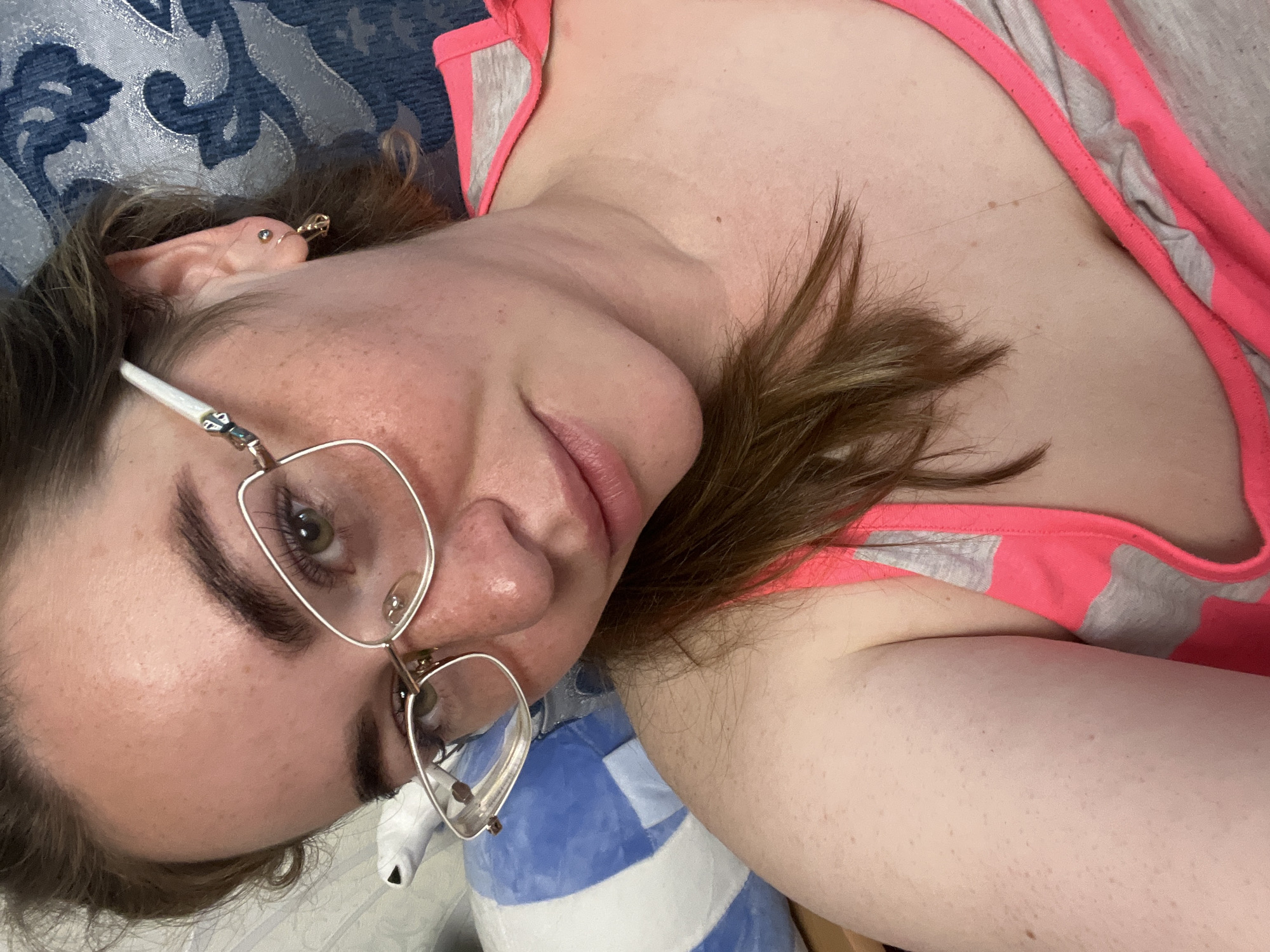 A close-up, slightly tilted portrait shows a person, likely female, lying down indoors. The subject has fair skin with visible freckles across their cheeks and nose, and light brown hair. They are wearing clear-framed glasses with thin metallic rims. Their left eye, visible through the glasses, is a light color, possibly green or hazel, and they are looking directly at the camera with a calm, relaxed expression, featuring a slight smile on their lips. The left ear is visible, adorned with a small stud piercing and a gold-colored hoop with a small charm.
The person is wearing a pink and grey tank top or camisole. In the background, to the upper left, is a dark blue fabric with a lighter blue ornate pattern, possibly a blanket or upholstered surface. Beneath the subject's chin and neck area, a blue and white patterned object, resembling a pillow or stuffed animal, is partially visible. The lighting is soft and even, suggesting an indoor setting without indicating a specific time of day. No text is visible in the image. While the provided context indicates Chaikovsky, Russia, there are no visual cues within the image itself to confirm this location.