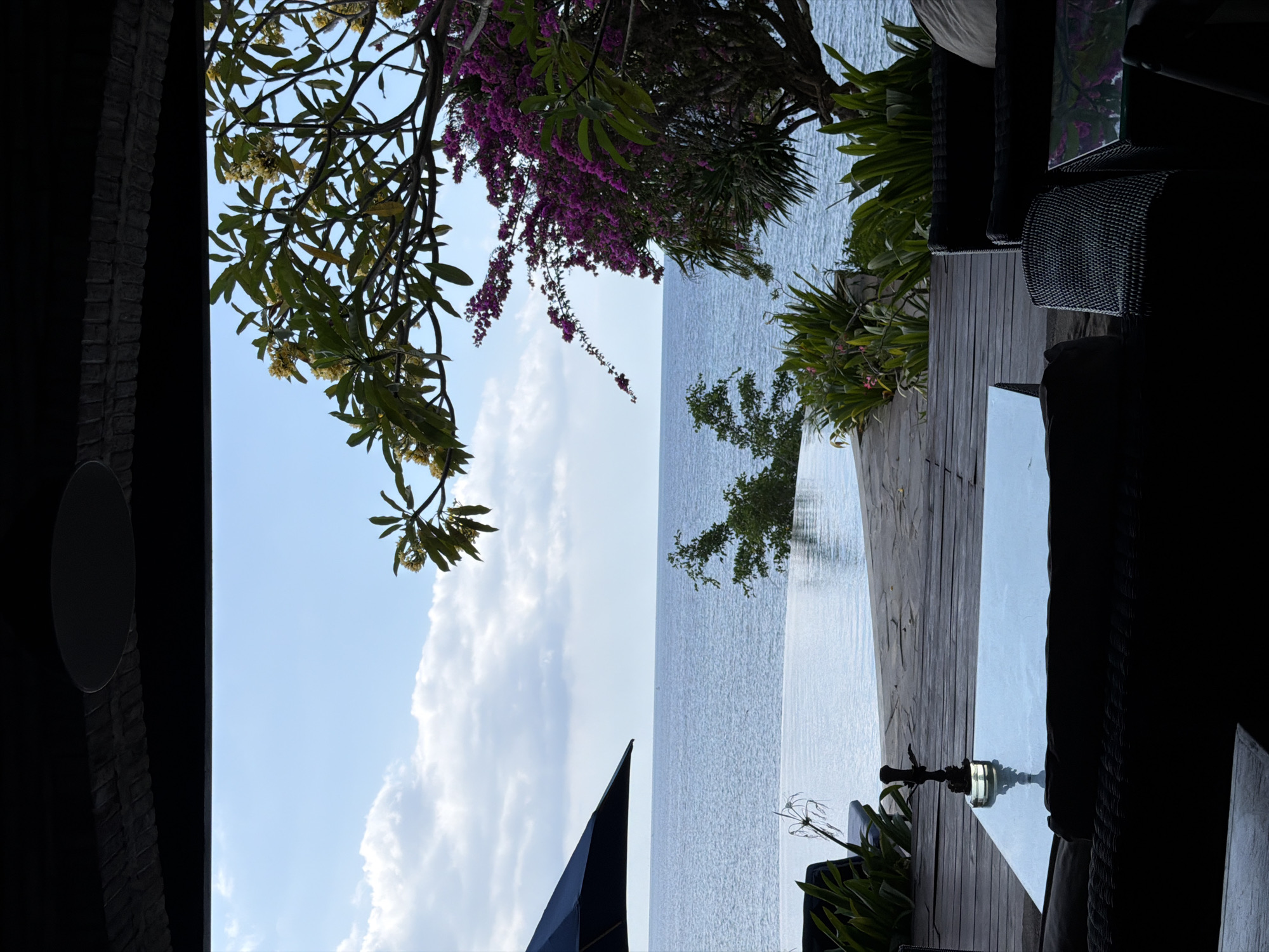 This is a photograph of an outdoor seating area with a view of the ocean in Gerokgak, Indonesia. In the foreground, there is a dark, silhouetted lounge area with wicker furniture and a glass-topped coffee table. On the coffee table sits a dark, metallic hookah. Beyond the seating area, a wooden deck leads to an infinity pool that appears to merge with the sea. Lush green and purple flowering plants frame the view of the calm, blue ocean under a bright, cloudy sky. A large black umbrella is visible to the left of the pool. The overall scene suggests a relaxed, tropical atmosphere, likely a resort or private villa.