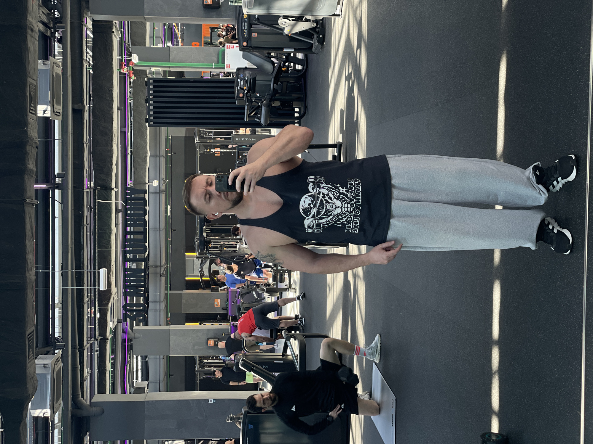 A man is taking a selfie in a gym. He is wearing a black tank top and gray sweatpants. Other people are exercising in the background. The location is Kazan, Russia.