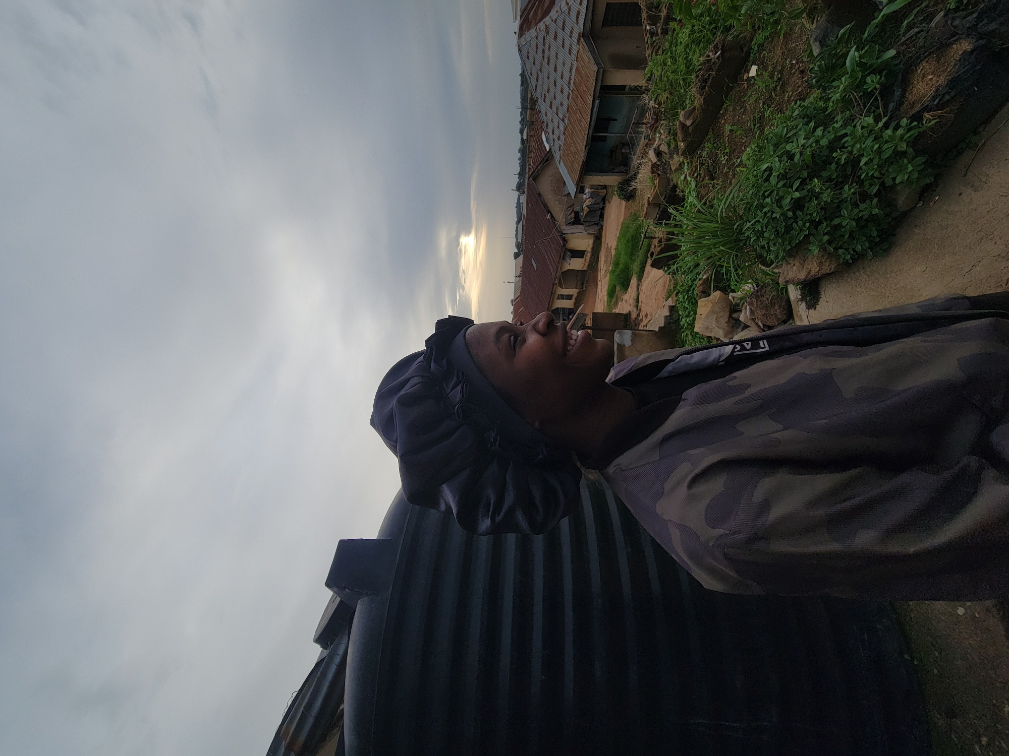 In Jos, Nigeria, a young woman is captured in a profile shot, her face turned to the left and illuminated by soft, natural light. She is wearing a dark bonnet and a camouflage jacket. Her expression is one of genuine joy, with a wide smile revealing her teeth and eyes crinkled at the corners. Behind her, a large black water tank dominates the left side of the frame. The background opens up to a view of a residential area with tin-roofed houses, some appearing weathered. The sky above is overcast with a hint of sunlight breaking through the clouds on the horizon. The foreground shows an uneven terrain with patches of green vegetation and scattered debris, suggesting an unpaved or underdeveloped area.